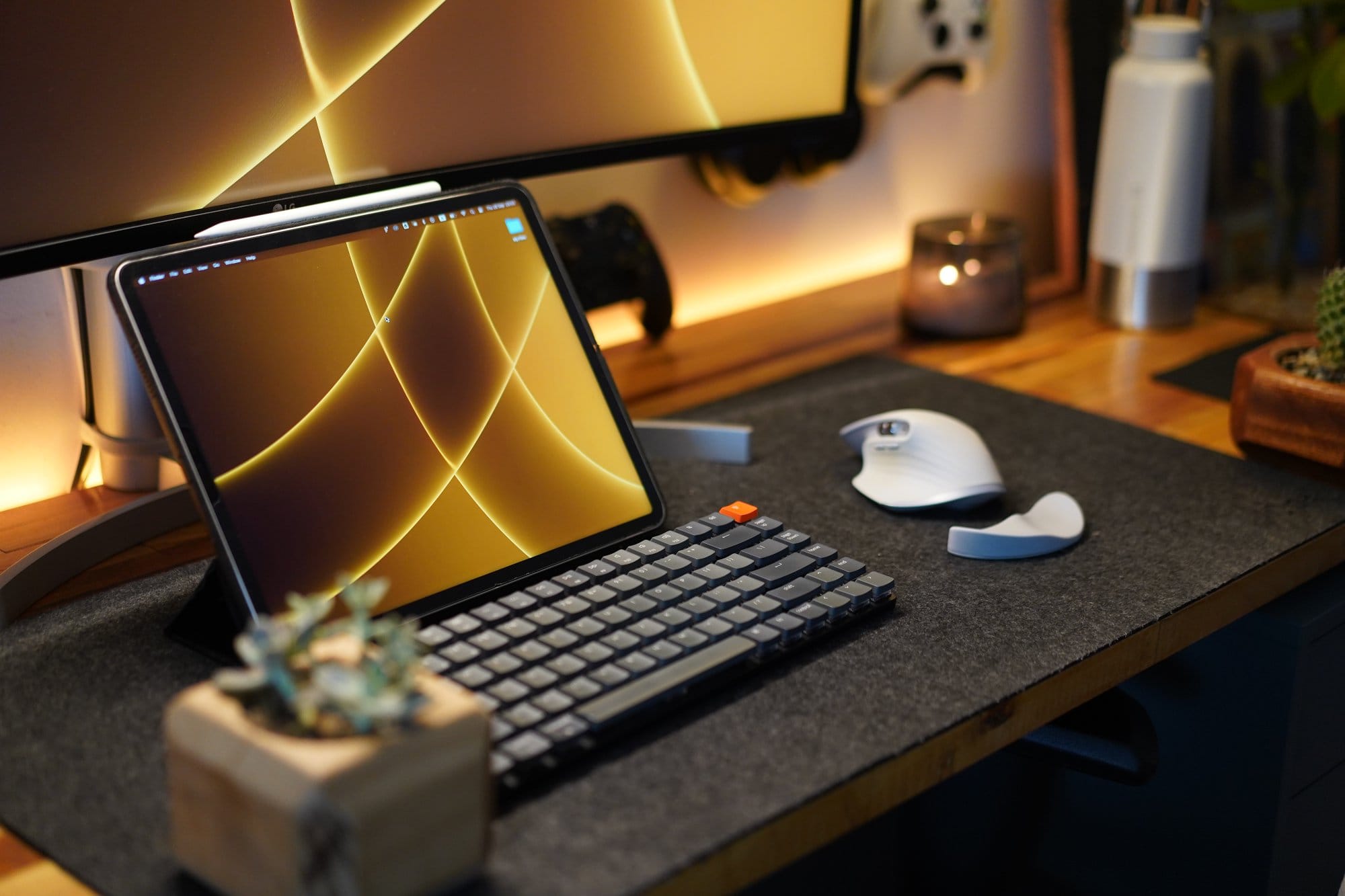 A workspace showing a tablet with a golden geometric wallpaper, a compact mechanical keyboard, a white ergonomic mouse, a felt desk mat, and a small potted succulent, set against warm ambient lighting