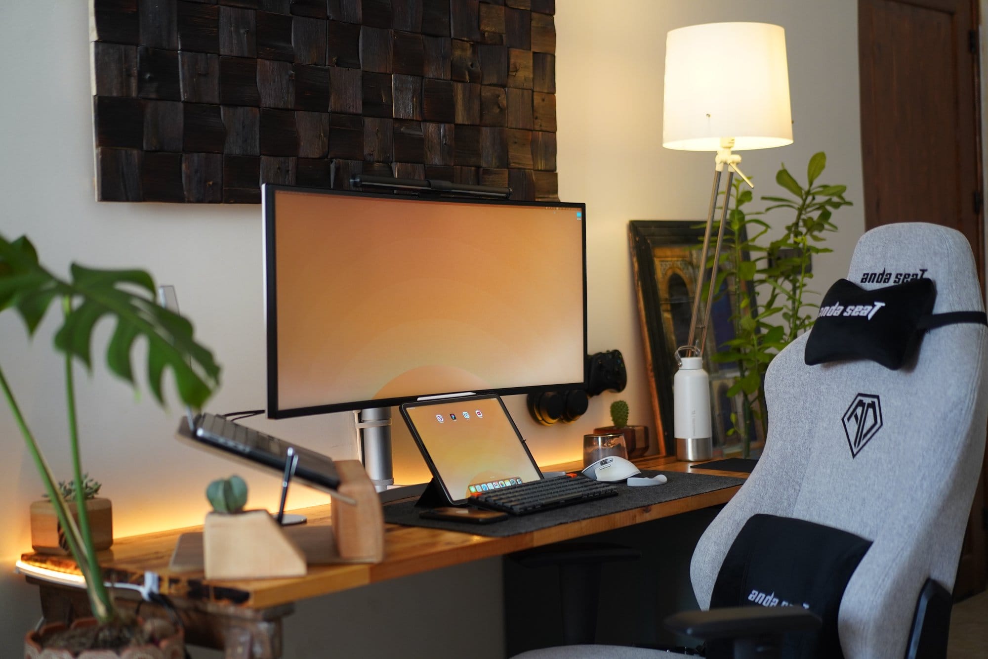 A workspace setup with a grey Anda Seat ergonomic chair, a wide monitor mounted on a wooden desk, a tablet and keyboard, surrounded by plants, a wooden laptop stand, and warm lighting accents