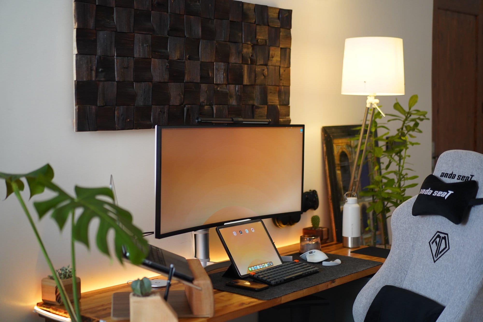 A modern workspace with an Anda Seat grey gaming chair, a wide LG monitor mounted on a wooden desk, an iPad and Keychron keyboard on a desk mat, and a floor lamp illuminating plants and decor in the background