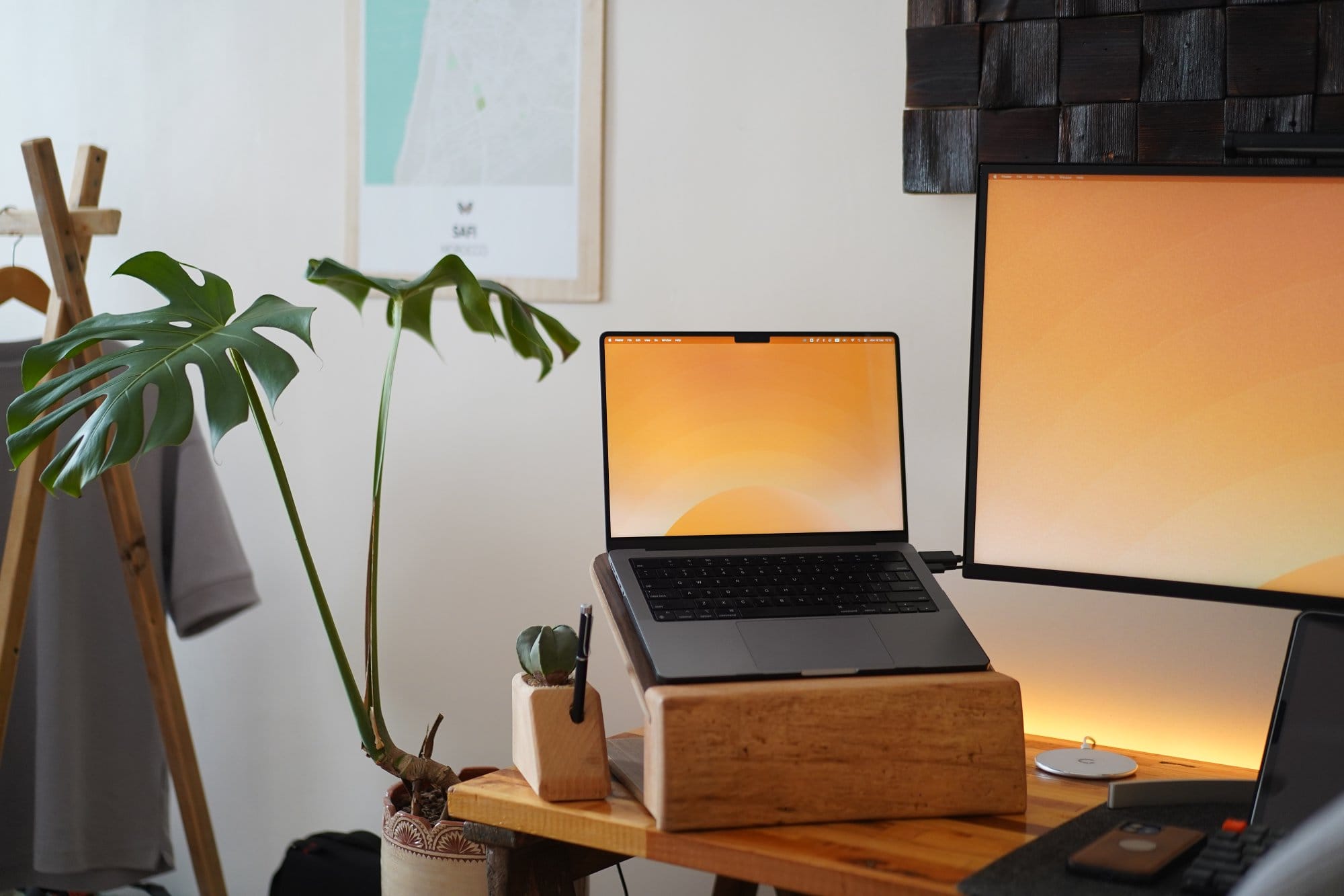 A 2021 Apple MacBook M1 Pro Max on a wooden laptop stand, placed beside a potted Monstera plant and a pen holder, with a minimalist desktop background matching the adjacent monitor