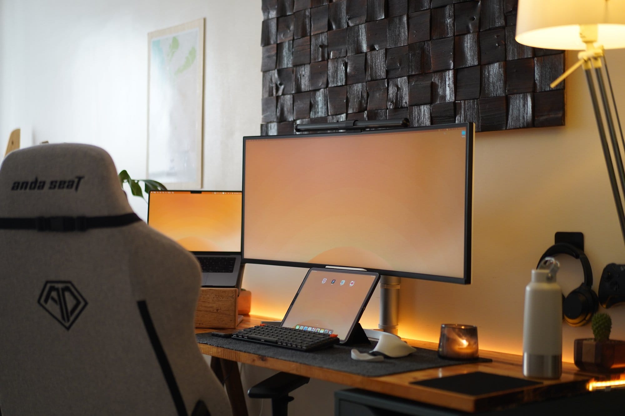 A grey Anda Seat gaming chair facing a desk setup with a wide LG monitor, a MacBook, and an iPad on a wooden desk, accompanied by ambient lighting and accessories like headphones and a water bottle