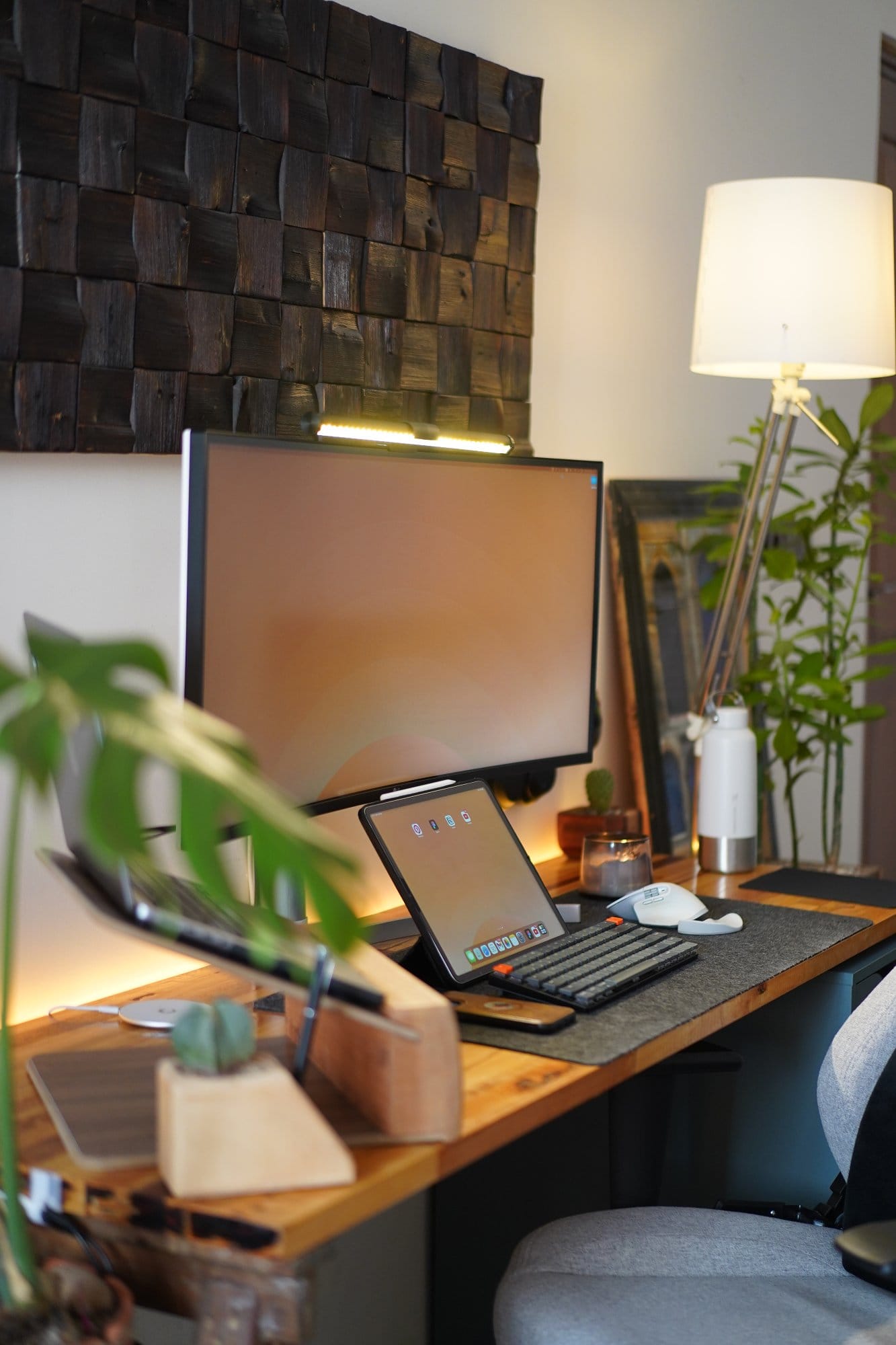 A desk setup with an LG 38WN95C-W monitor lit by an Eye-Care Desk Lamp, a 2021 Apple 12.9-inch iPad Pro paired with a Keychron K3 keyboard, a Logitech MX Master 3S mouse, and surrounded by plants and warm ambient lighting