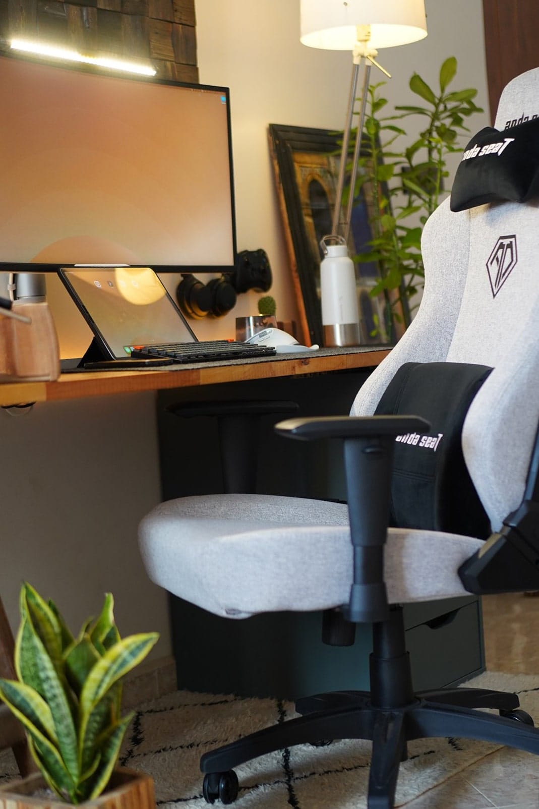 A grey Anda Seat Phantom 3 chair positioned next to a wooden desk setup with a monitor, tablet, and keyboard, with a snake plant in a wooden planter on a patterned rug below