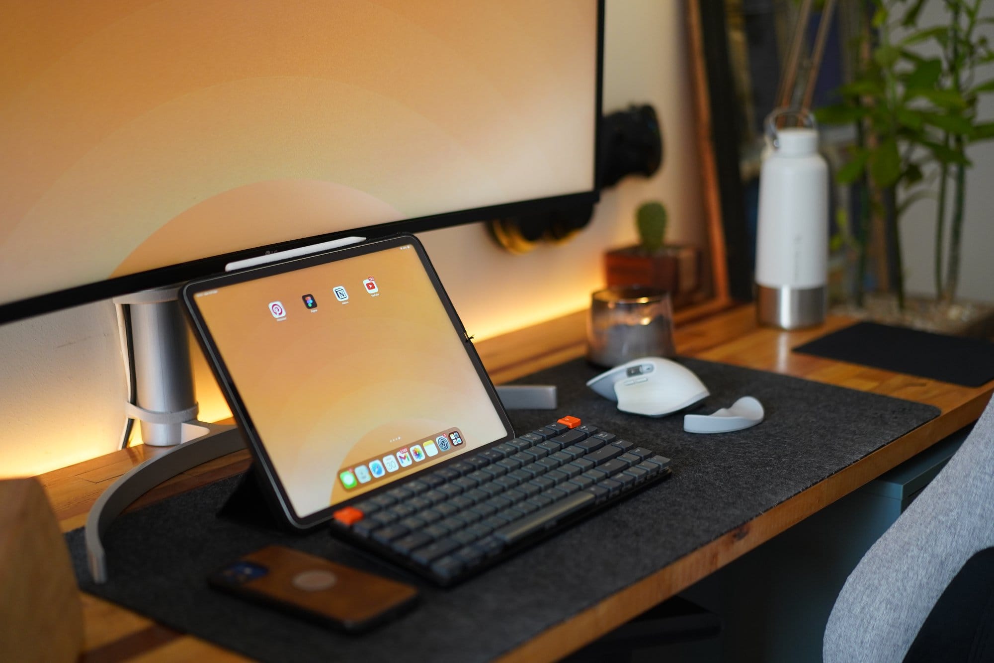 An iPad rests on a desk with a Keychron keyboard, Logitech mouse, and a leather phone case, surrounded by a monitor, candle, and plants