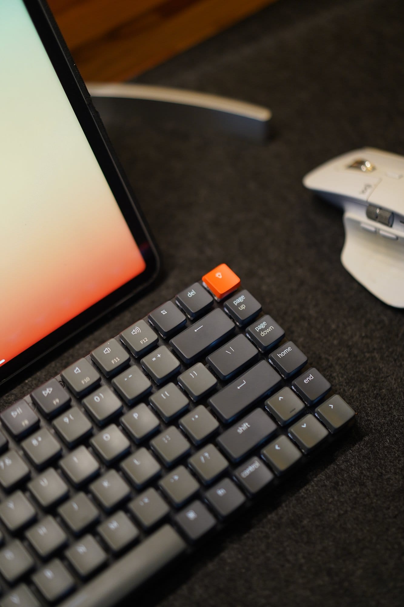 A close-up of a Keychron keyboard with an orange escape key, next to an iPad and a Logitech mouse on a felt desk mat