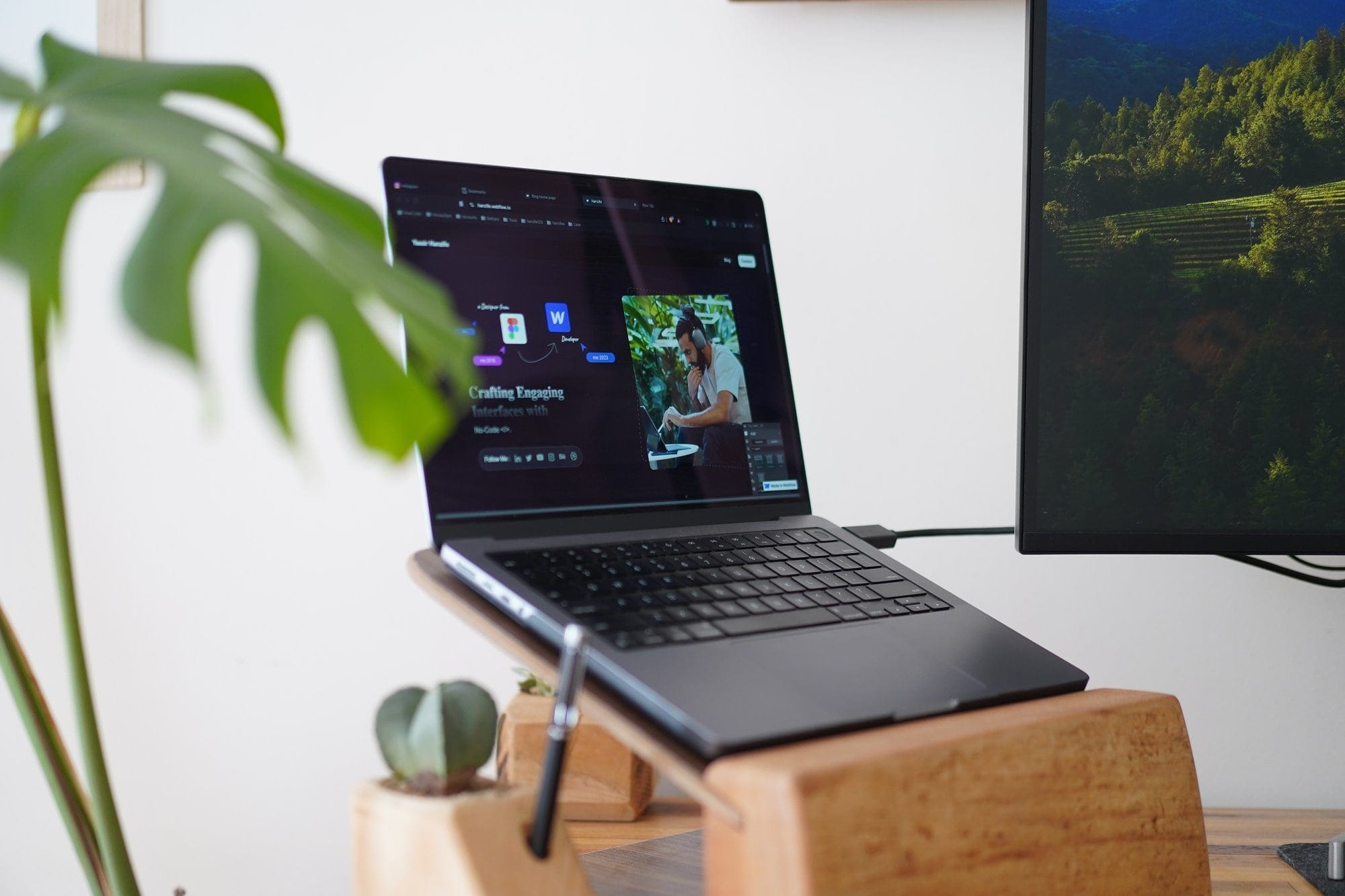 A 2021 Apple MacBook M1 Pro Max displaying a web design workflow, placed on a wooden laptop stand next to a small potted cactus and a pen holder, with a Monstera plant in the foreground