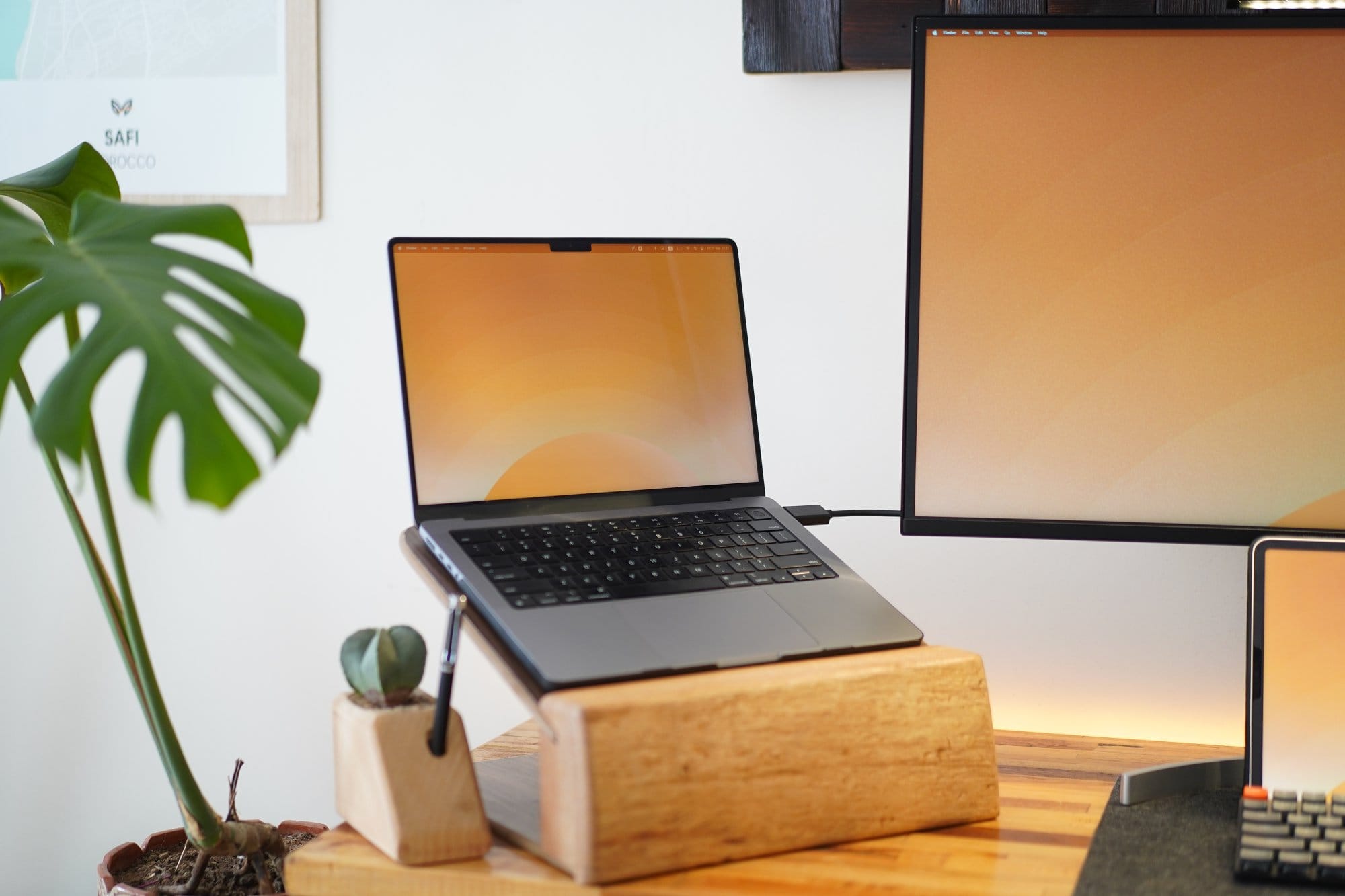 A laptop sits on a wooden stand next to a monitor, with a small potted plant and pen holder on the desk