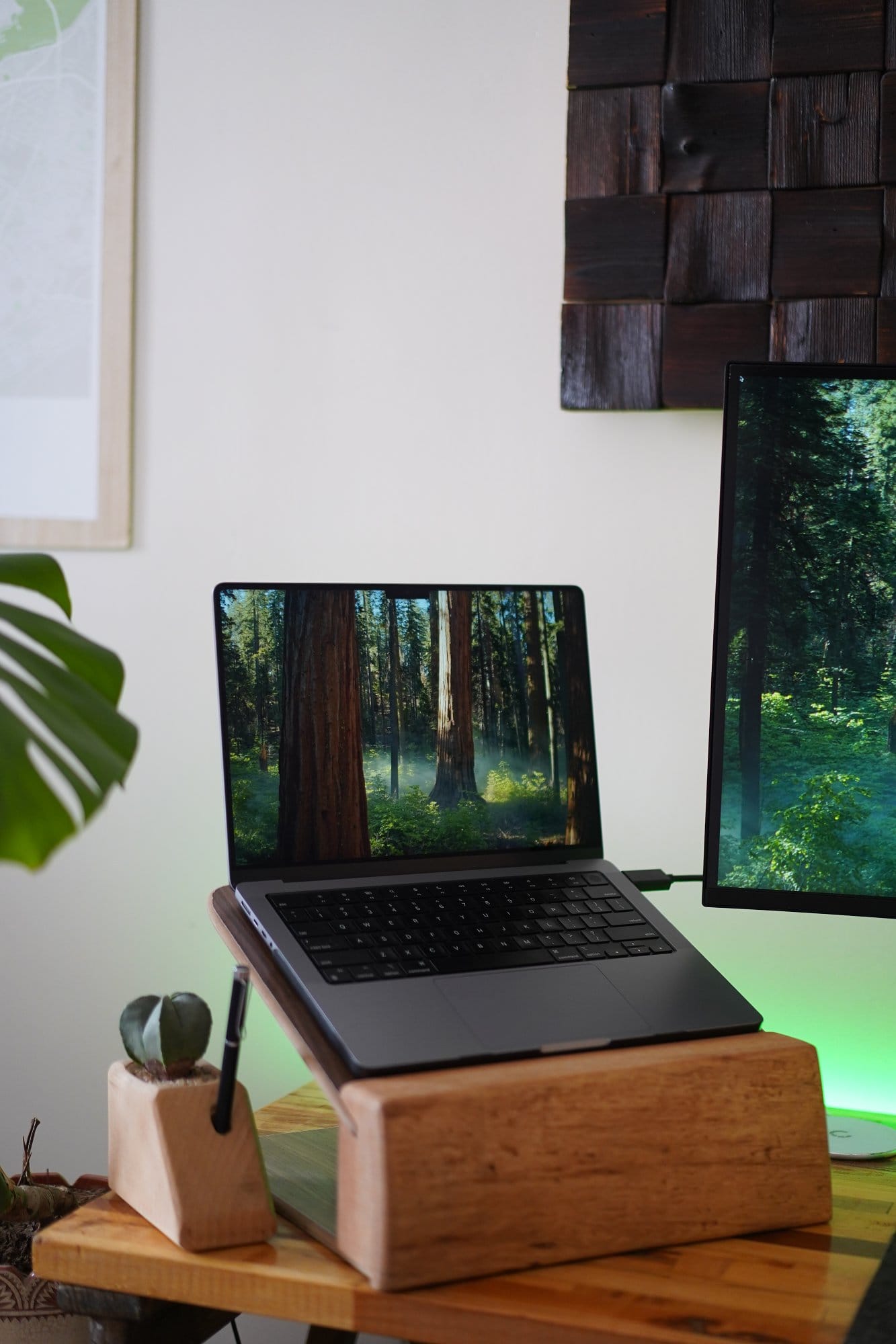 A 2021 Apple MacBook M1 Pro Max displayed on a wooden laptop stand, next to a small plant and pen holder, with a forest-themed desktop background