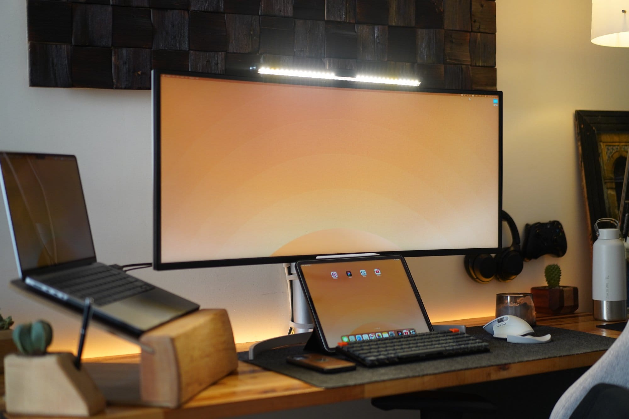 A wide curved monitor with a light bar sits above an iPad Pro, a mechanical keyboard, and a mouse on a desk mat, with a laptop elevated on a wooden stand to the left