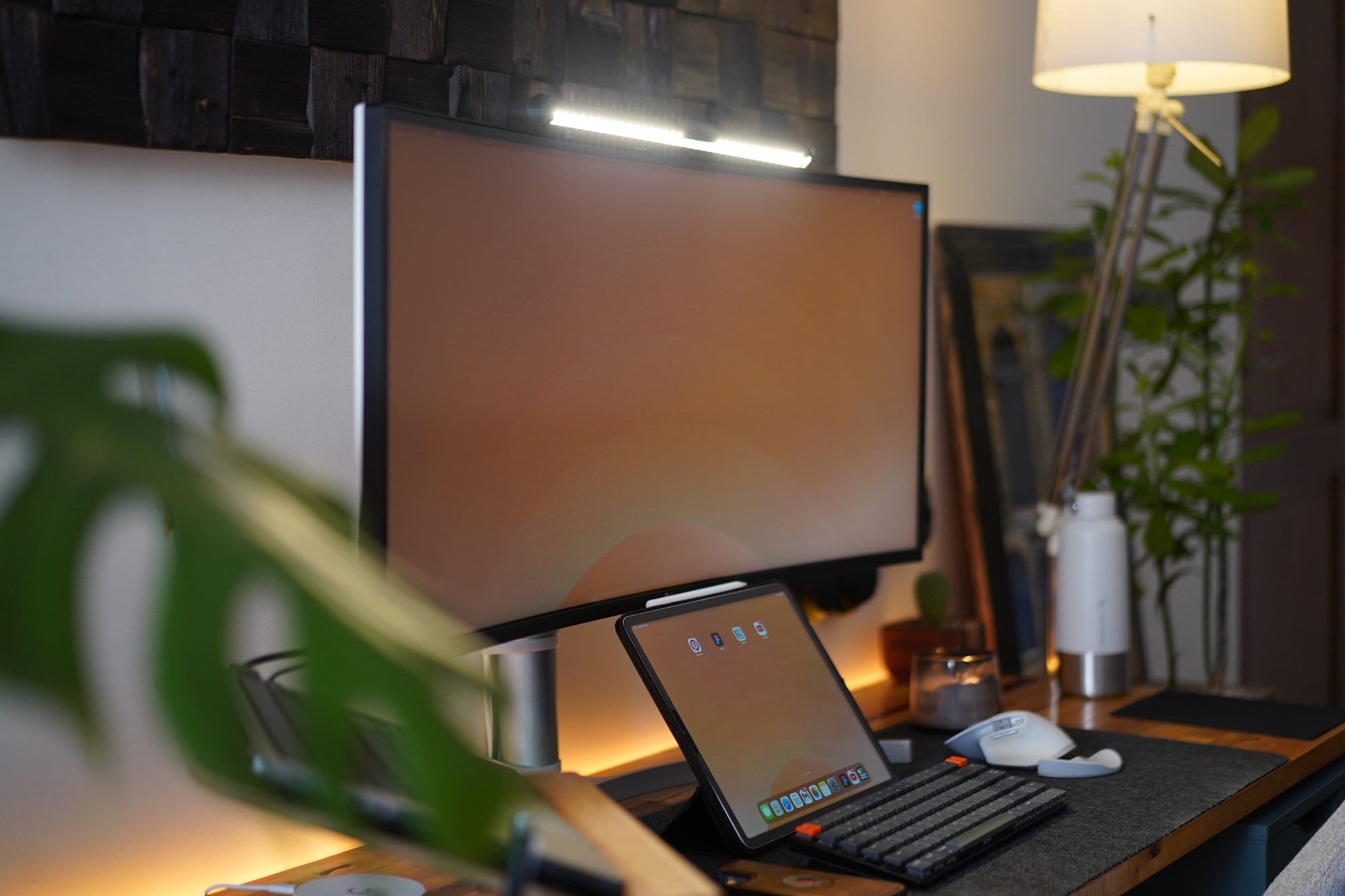 A workspace setup with a curved monitor displaying a muted orange background, a tablet with matching wallpaper, a compact mechanical keyboard, a white ergonomic mouse, and a leafy plant partially in the foreground, all illuminated by soft, warm lighting