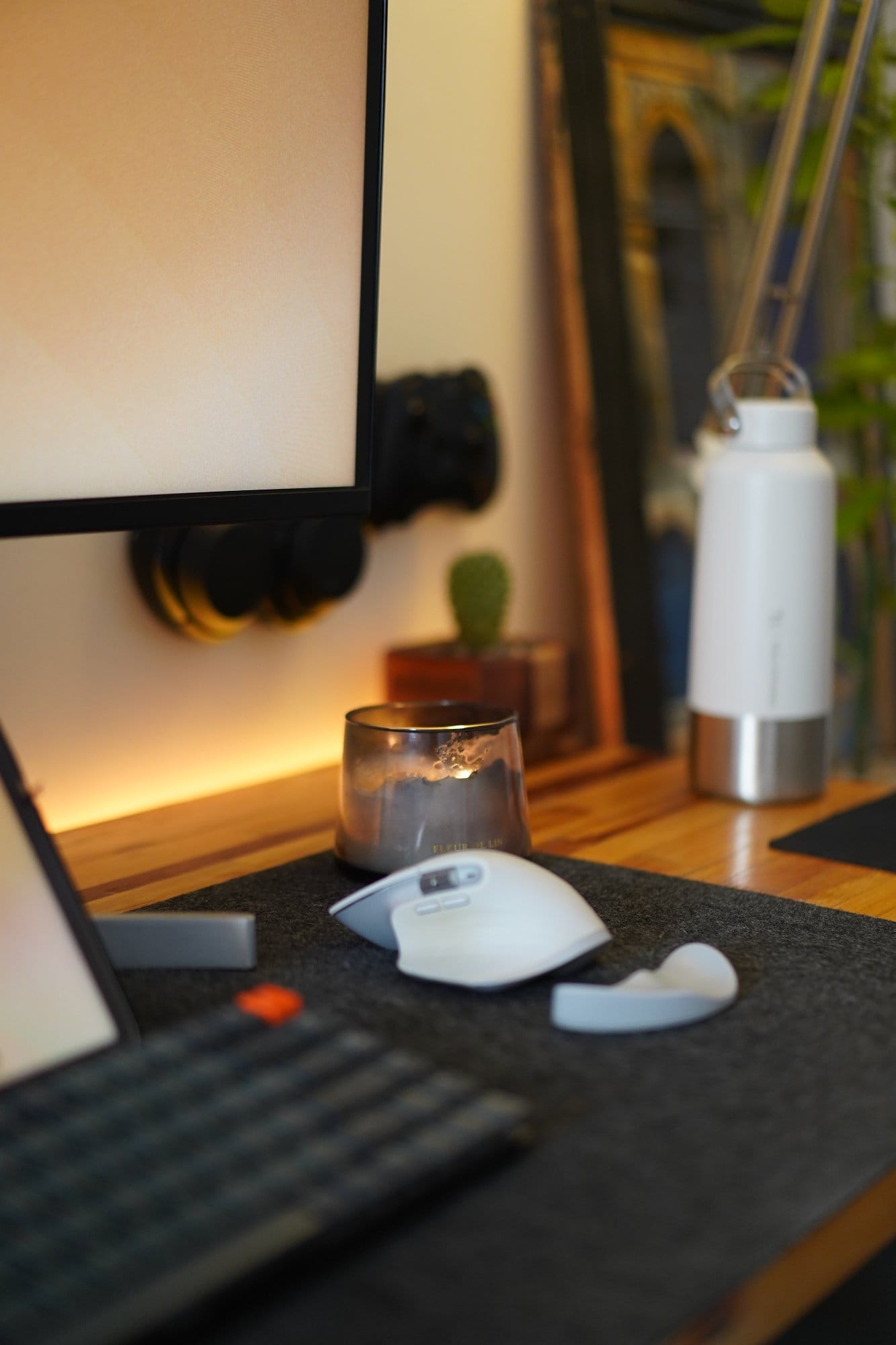 A close-up of a Logitech MX Master 3S mouse on a felt desk mat, with a grey glass candle, a small cactus in a wooden pot, and a white insulated bottle in the background