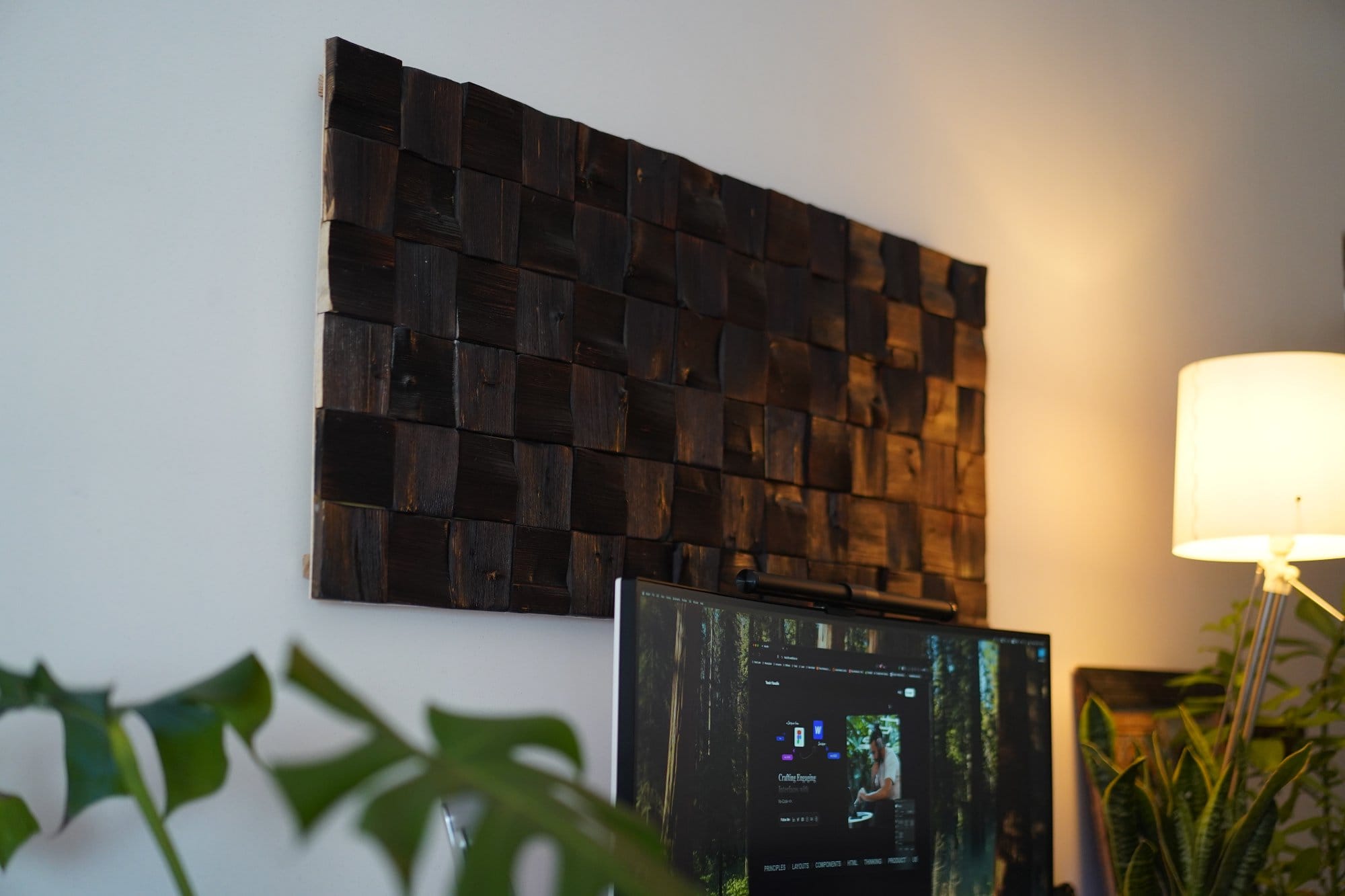 A wall-mounted wooden acoustic panel hangs above a monitor, accompanied by a floor lamp and some greenery in the background