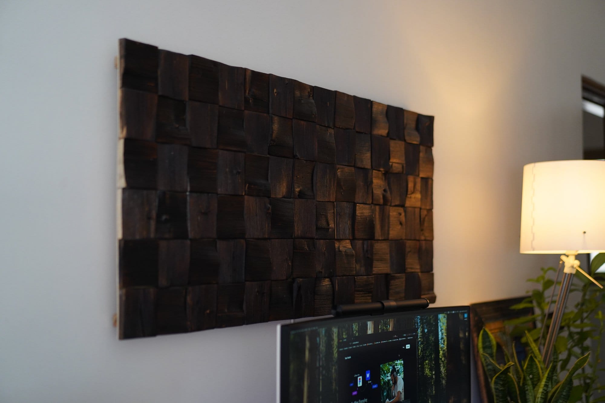  A dark wooden wall panel with a woven texture hangs above a desk setup, partially visible with a monitor below and a lamp to the side