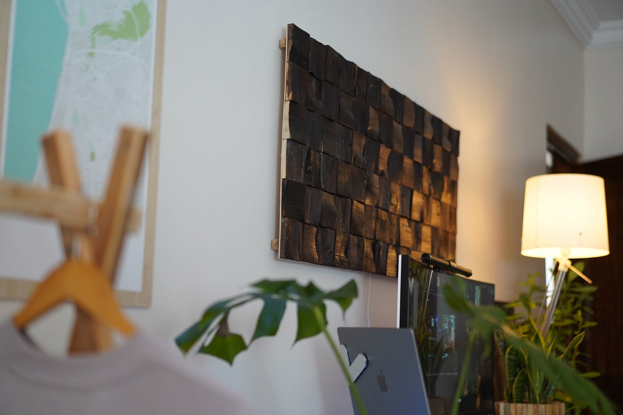 A close-up of a dark wooden wall panel above a desk setup, with a standing lamp and a glimpse of a laptop and greenery in the foreground