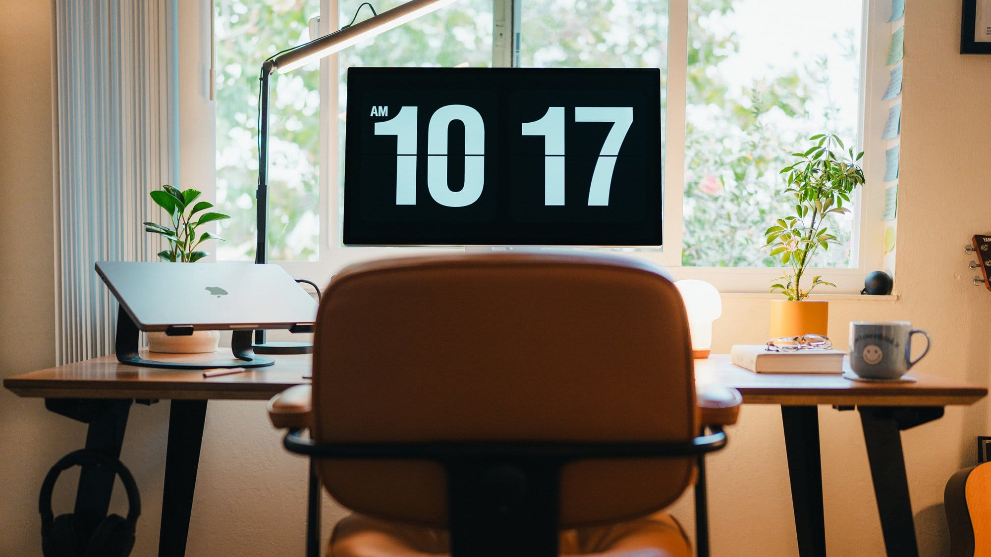 A workspace setup with an Apple Studio Display showing a digital clock, a MacBook on a laptop stand, a desk lamp, potted plants, and a mug on the desk, framed by a large window