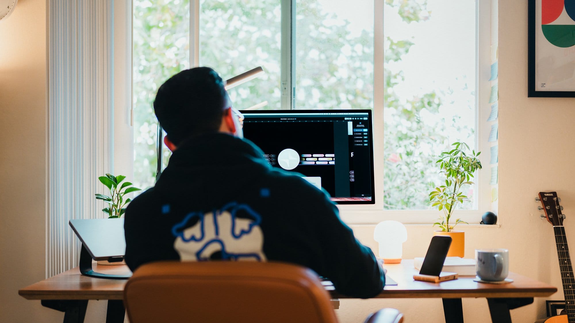 A person sitting on an IKEA ALEFJÄLL chair, working at a Floyd table with an Apple Studio Display and a Twelve South Curve laptop stand, framed by plants and a guitar in the background