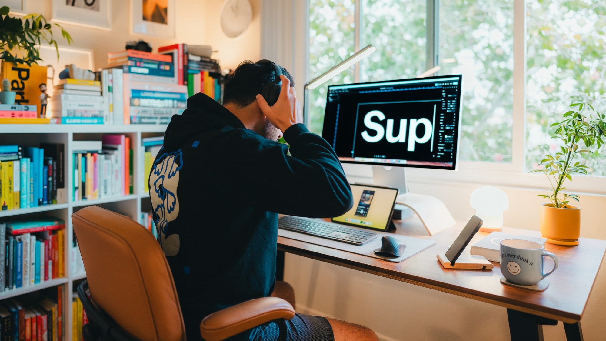 A person wearing headphones sits at a wooden desk with an Apple Studio Display showing the text “Sup,” an iPad on a stand, and a mug with “don’t overthink it“ next to a small yellow plant pot