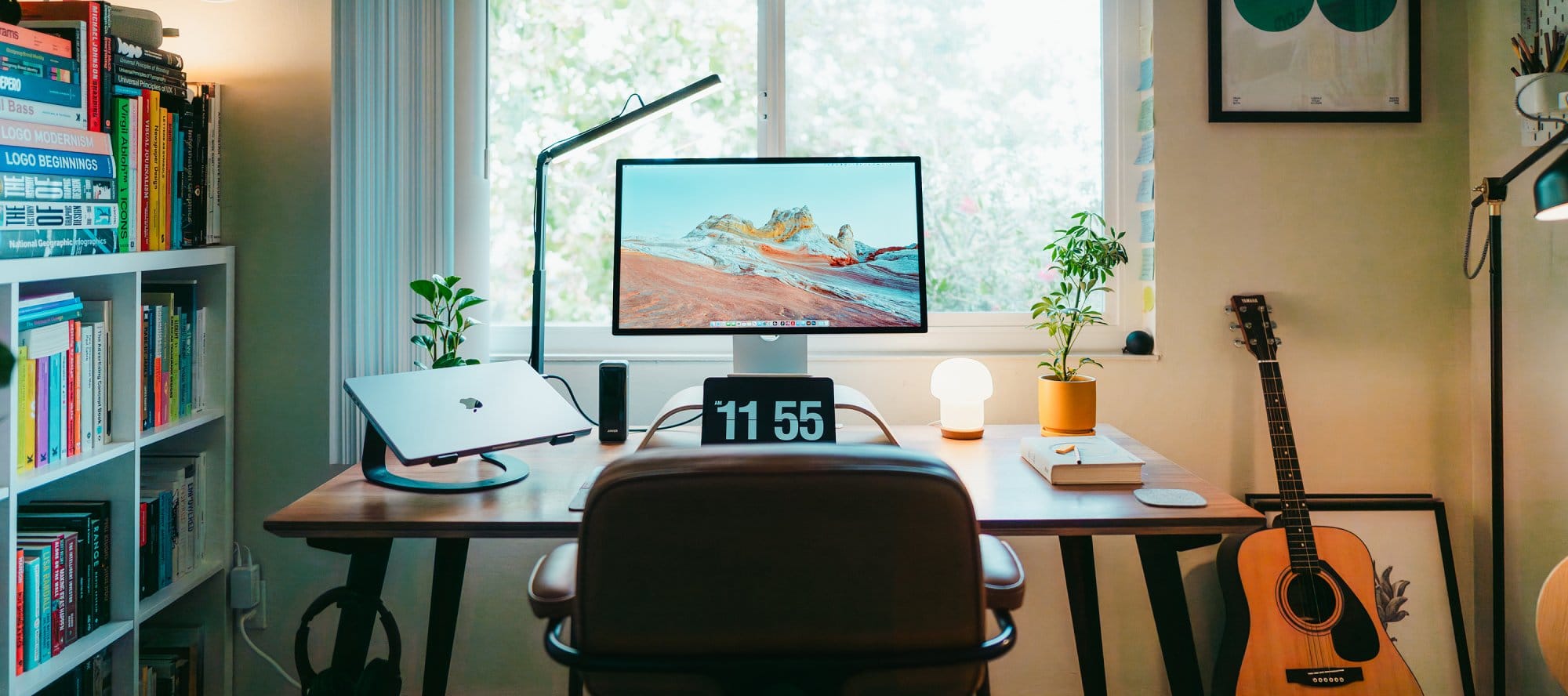 A workspace with an Apple Studio Display, a MacBook on a laptop stand, a Yamaha acoustic guitar leaning against the wall, and shelves filled with colourful books, lit by natural light from a large window