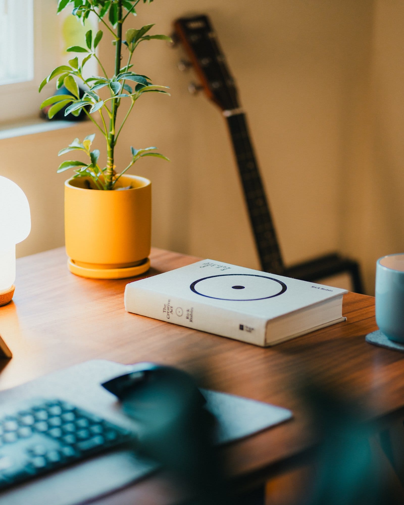 A wooden desk with a yellow plant pot, a book titled “The Creative Act: A Way of Being” by Rick Rubin, a lamp, and a guitar leaning against the wall in the background