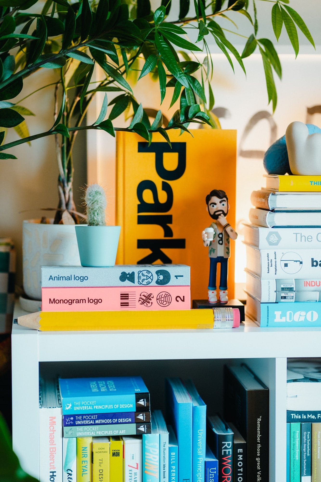 A close-up of a shelf holding books including “Park,“ “Animal Logo,“ and “Monogram Logo,“ a small potted cactus, a pencil-shaped decor item, and a figurine of a bearded man holding a cup