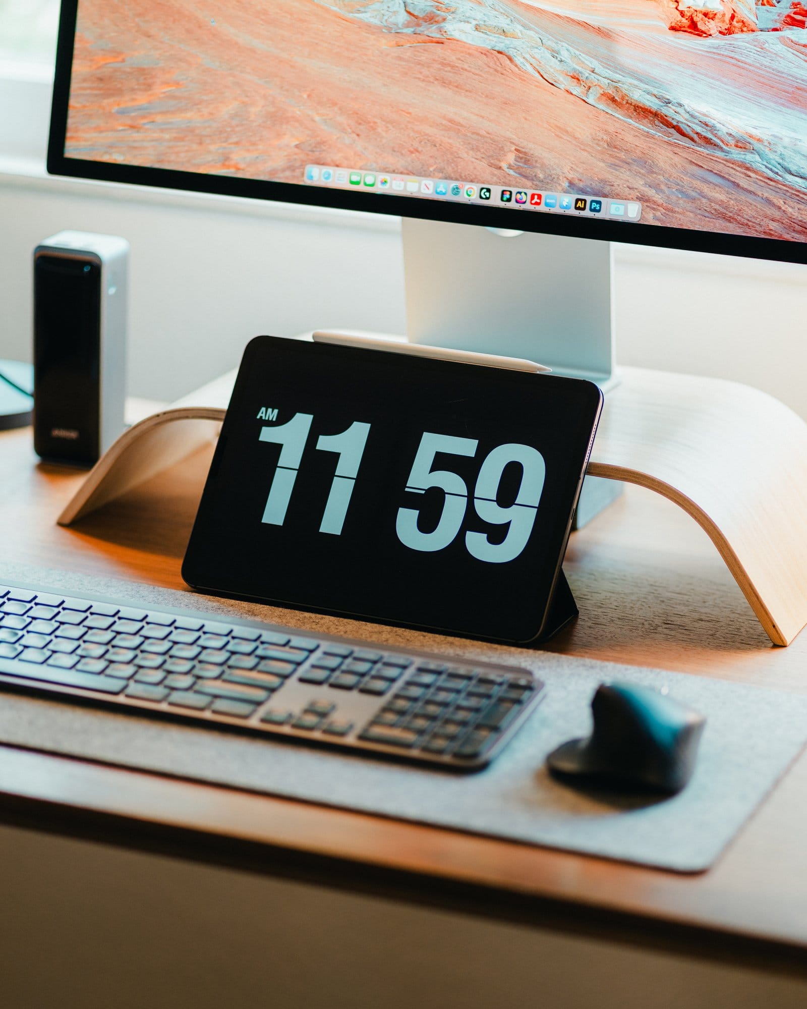 A close-up of a desk setup with a Logitech MX Keys keyboard, a Logitech MX Master 3 mouse, a tablet displaying the time “11:59 AM” on a wooden stand, and an Apple Studio Display showing a desert landscape