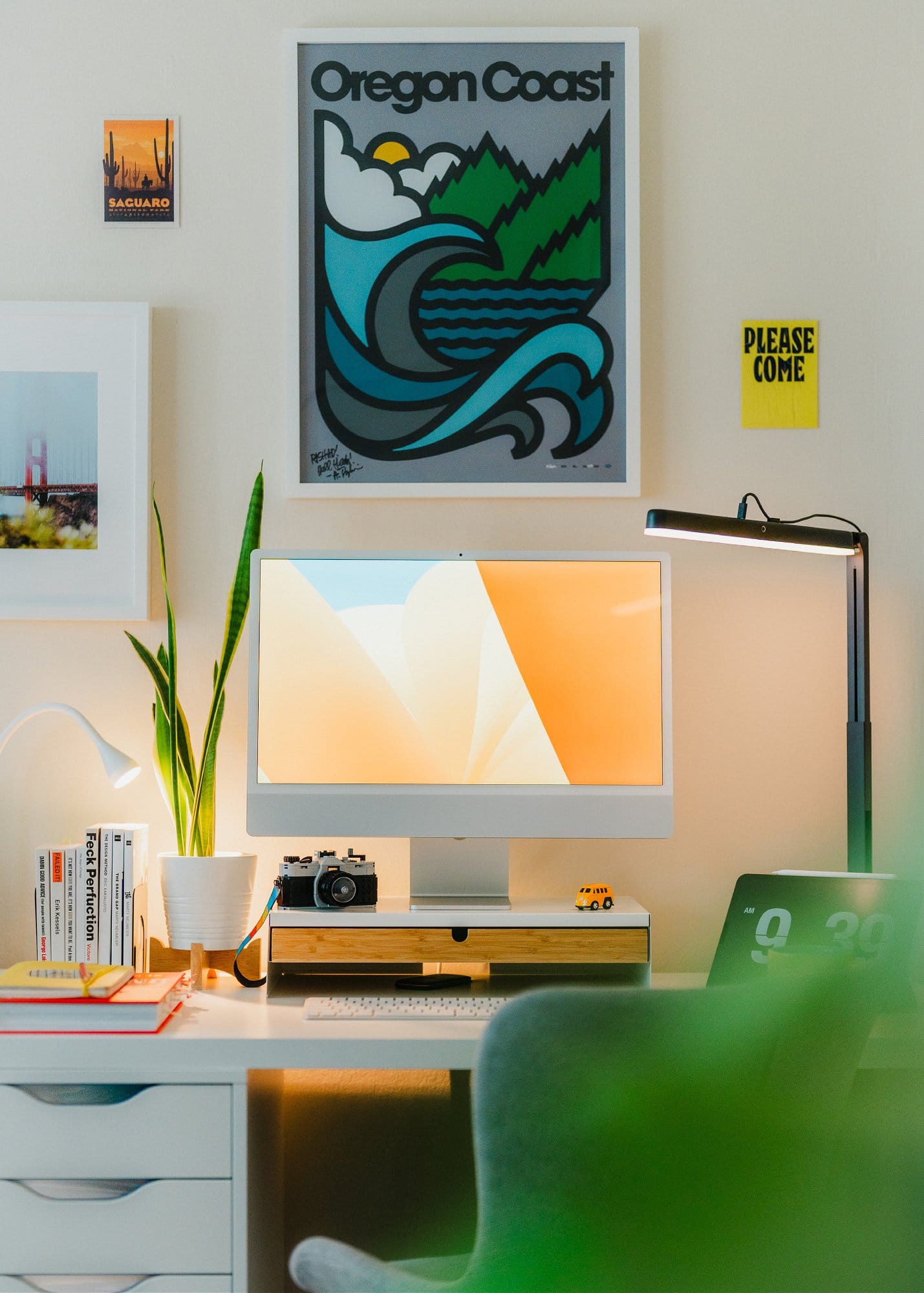 An Apple Studio Display on a Grovemade monitor stand, with a vintage camera, books, and plants on a white desk, illuminated by a desk lamp and surrounded by colourful wall art including an Oregon Coast poster