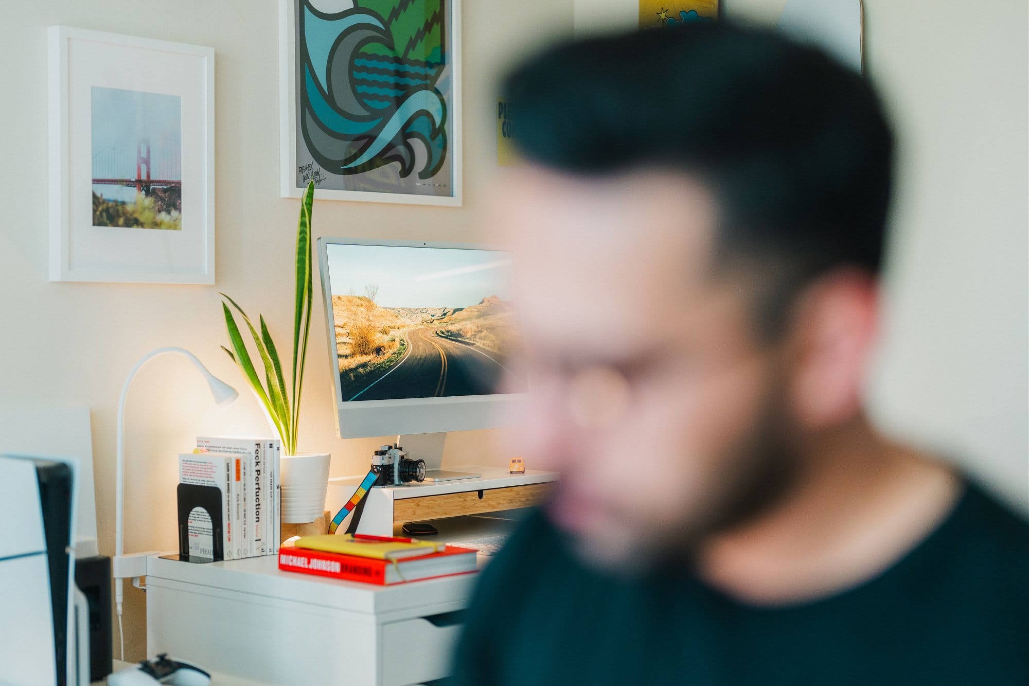 A workspace with an Apple Studio Display showing a road landscape, a snake plant in a white pot, and books including “Feck Perfuction“ on a desk, with a blurred figure in the foreground