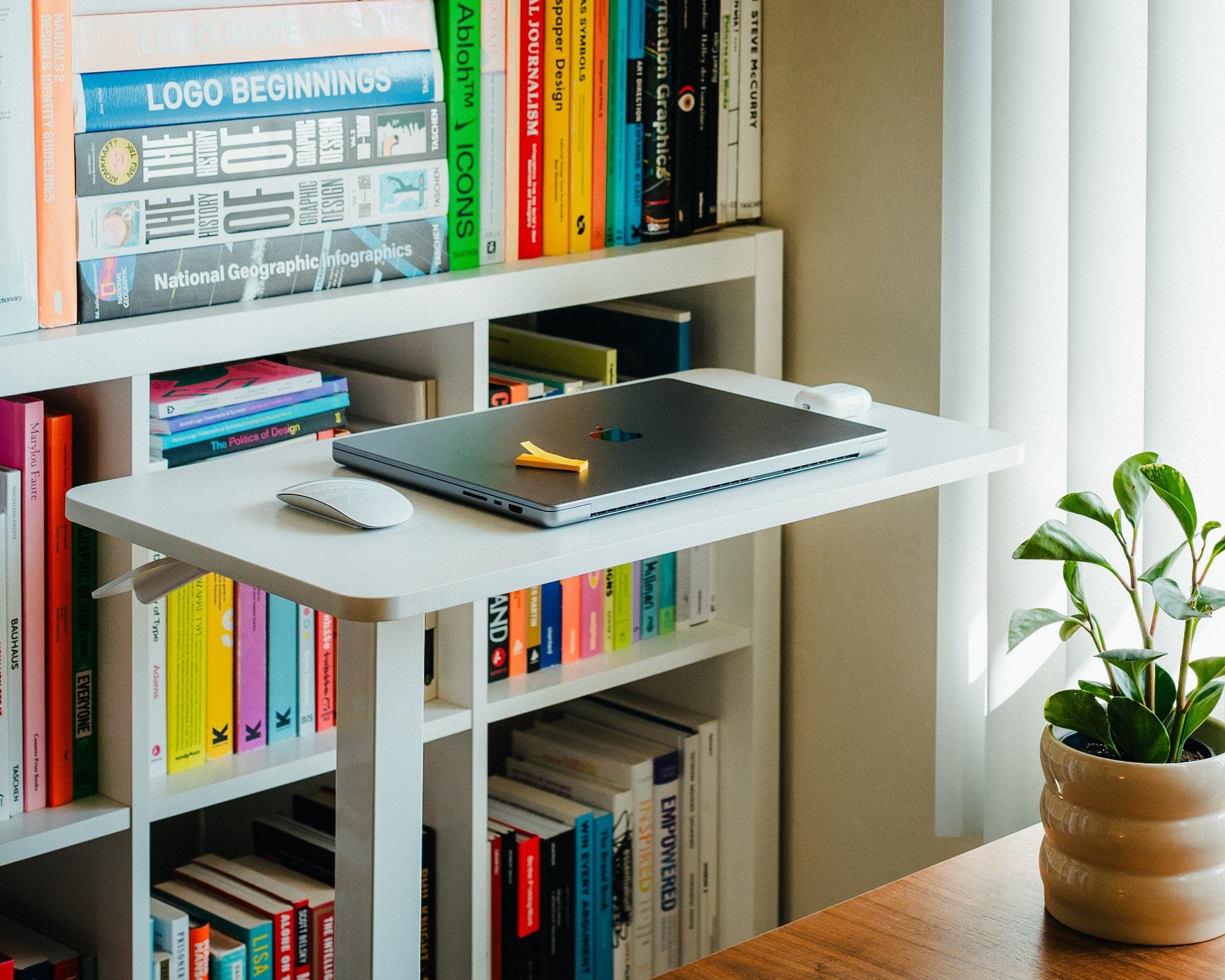 A white standing desk with a closed MacBook Pro 16” M1 and Magic Mouse placed in front of a colourful bookshelf filled with design and photography books, alongside a small green potted plant on the wooden table nearby