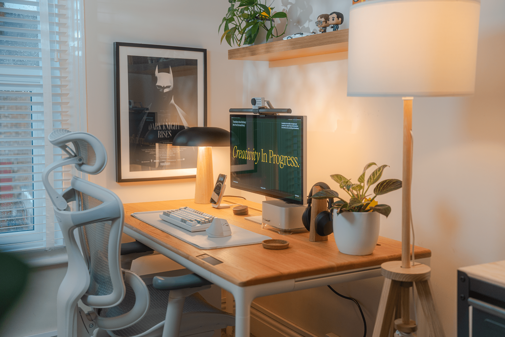 A modern desk setup with an Apple Studio Display, BenQ Halo monitor light bar, Keychron Q1 Max keyboard, Logitech Lift mouse, and Sony XM5 headphones, placed on a Beflo Tenon desk next to a white ergonomic chair and decorated with a potted plant and a “Dark Knight Rises” poster on the wall