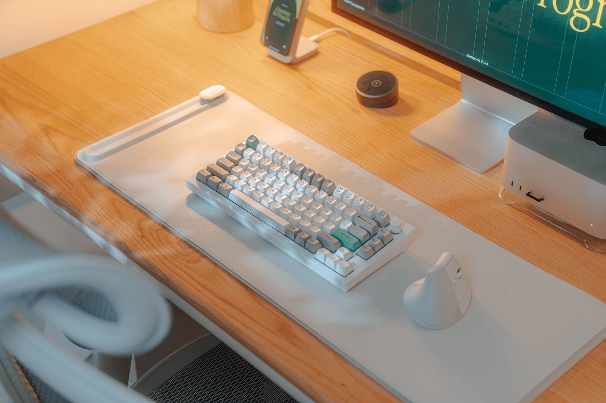 A wooden desk setup with a Keychron Q1 Max keyboard on an Orbitkey Slim desk mat, a Logitech Lift mouse, an Apple Mac Studio on a Spigen stand, and a smartphone on a Nomad Stand One MagSafe charger