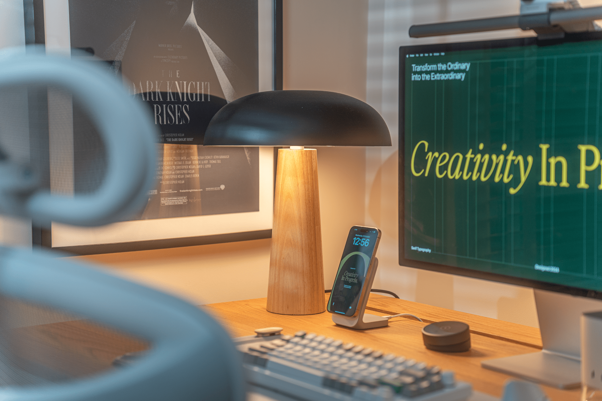 A close-up of a desk with a wooden lamp, a Nomad Stand One MagSafe charger holding a smartphone, and an Apple Studio Display showing “creativity in progress,” with a Batman poster by Olly Moss visible in the background