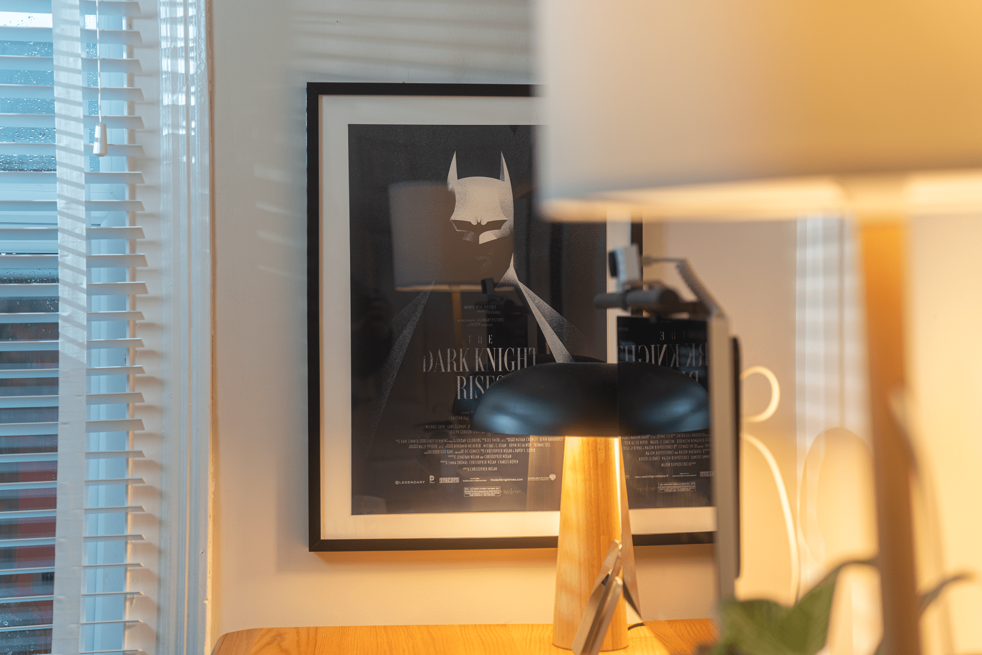 A framed “The Dark Knight Rises” poster is mounted on the wall near a wooden desk, with a modern lamp in the foreground and blinds partially covering a window to the left