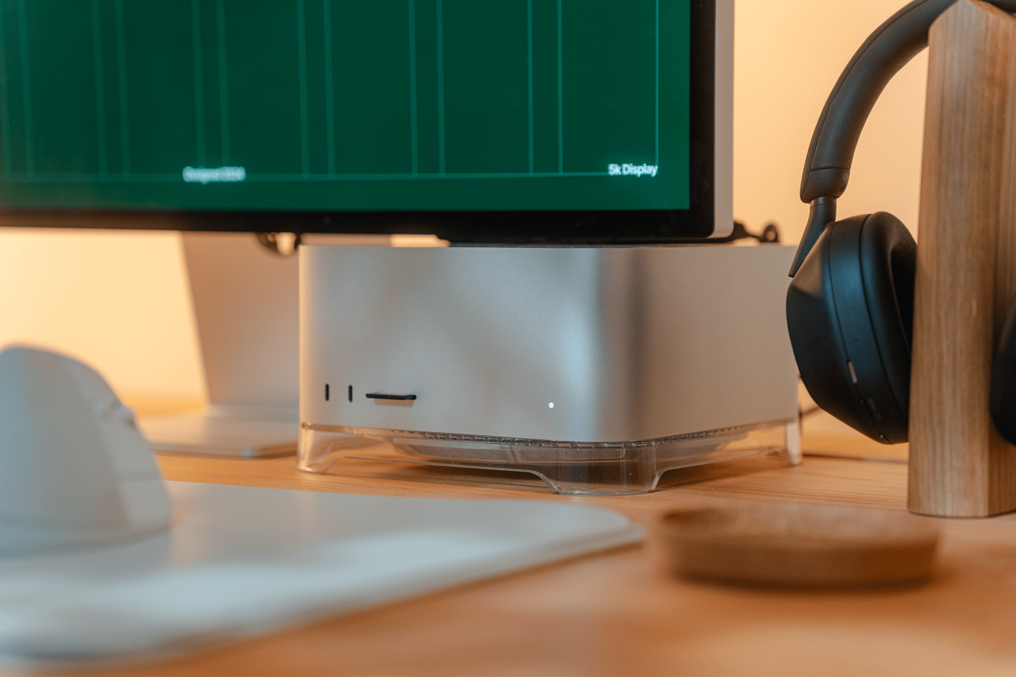 An Apple Mac Studio placed on a clear Spigen stand, with Sony XM5 headphones resting on a wooden stand beside it, and part of a Logitech Lift mouse visible on a desk mat in the foreground