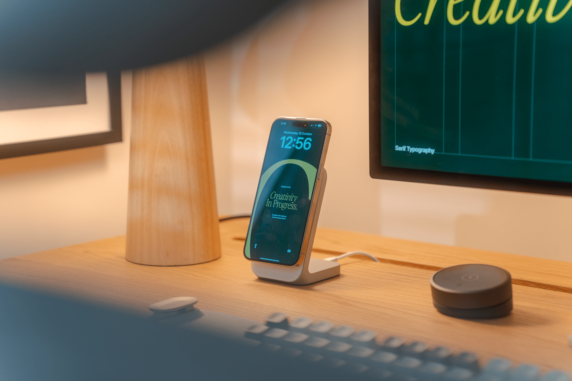 A smartphone displaying “Creativity in Progress” is propped on a charging stand on a wooden desk, alongside a lamp and a small smart speaker