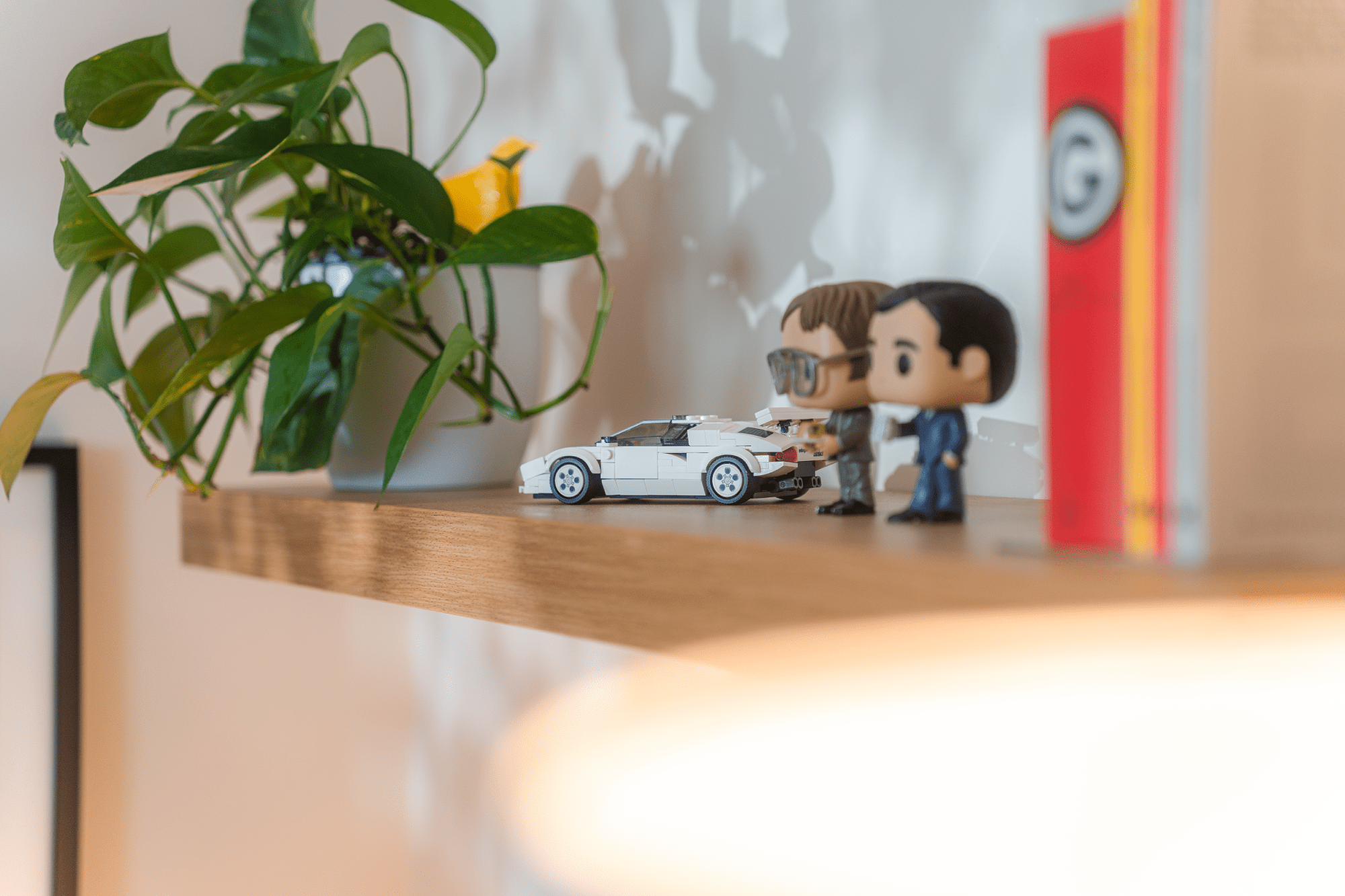 A wooden shelf featuring a small white model car, two collectible figurines, a leafy green plant in a ceramic pot, and a red book, all softly lit with warm light and subtle shadows