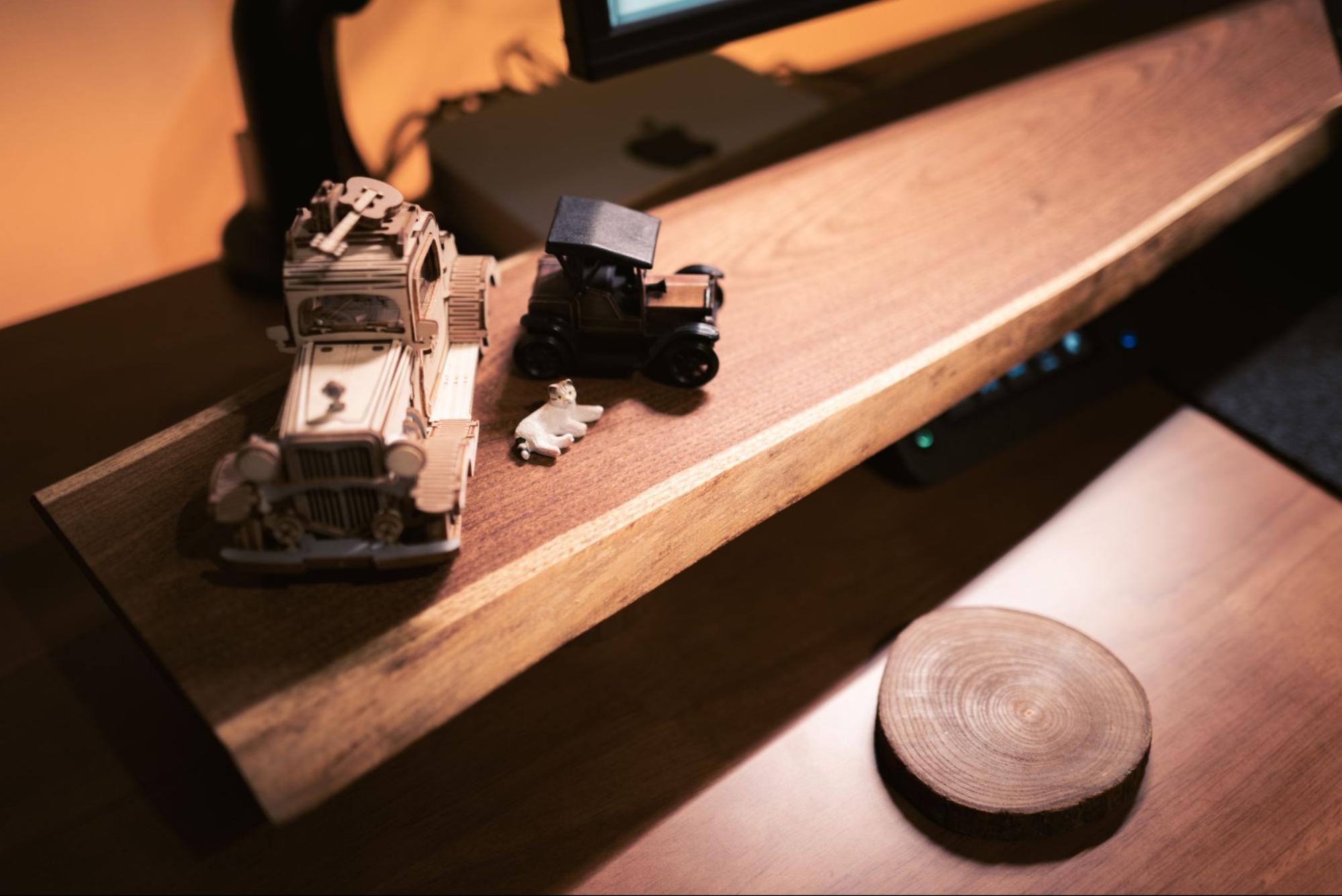 A close-up of a DIY desk shelf holding a wooden model truck, a small toy car, and a miniature figurine of a white animal, with a wooden coaster nearby