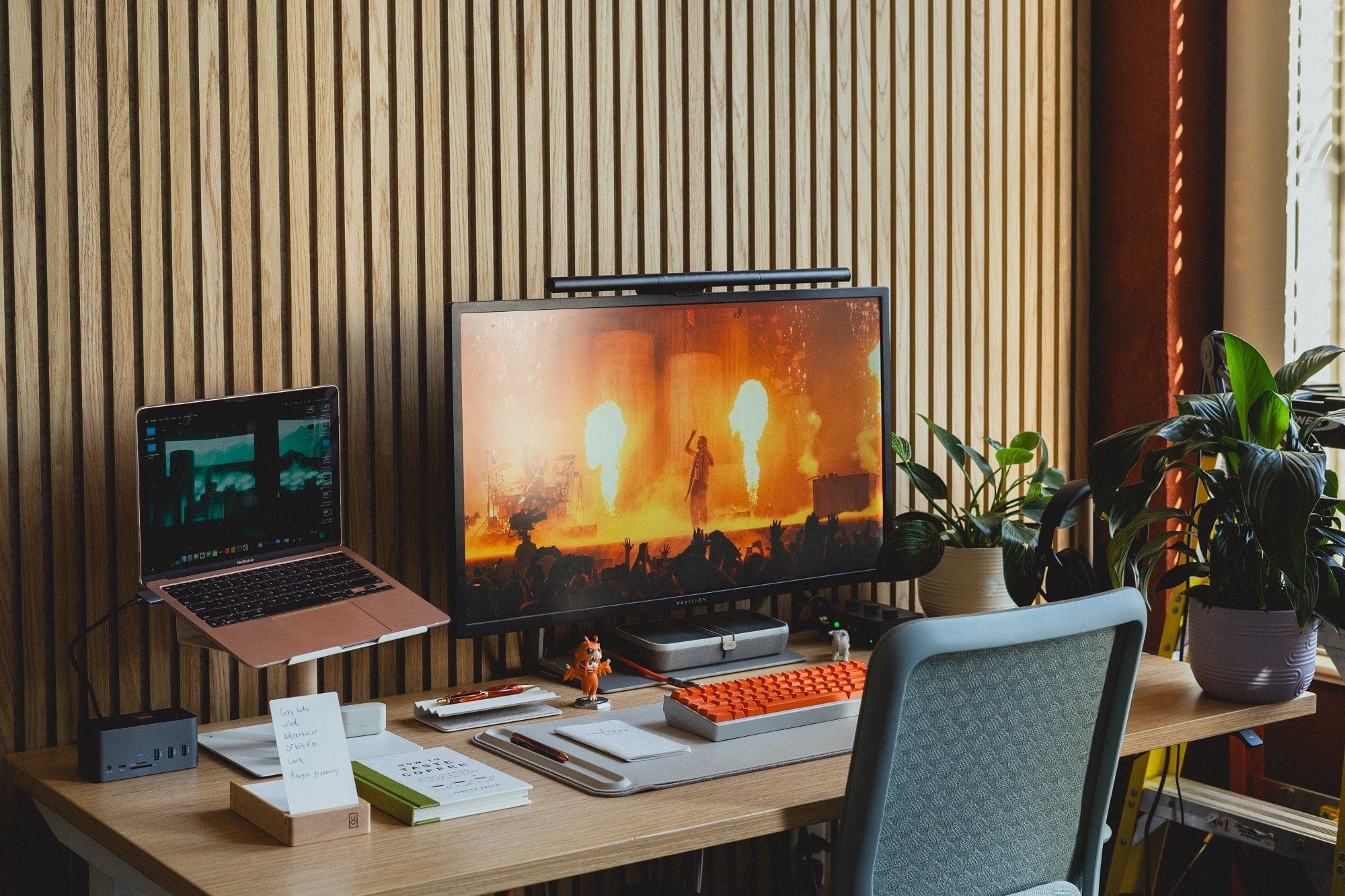 An M1 MacBook Air on an Ugmonk stand next to an HP Pavilion monitor displaying a concert, with a Branch Verve chair and plants around the desk