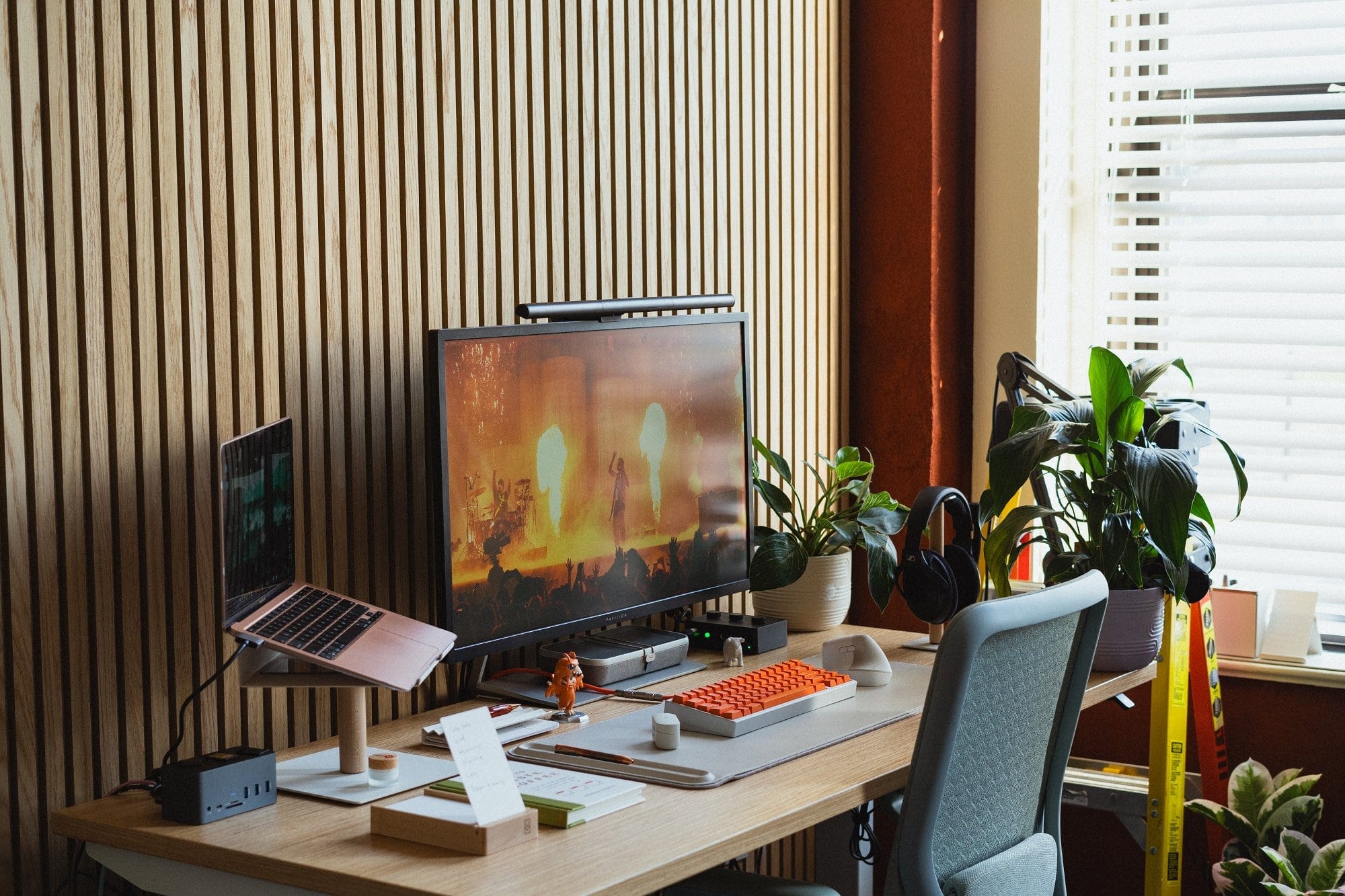 A home desk setup showing an M1 MacBook Air on an Ugmonk stand, a monitor playing a Twenty One Pilots concert, a KMG B65 keyboard with orange DCX Topaz keycaps, and various plants near a window with blinds