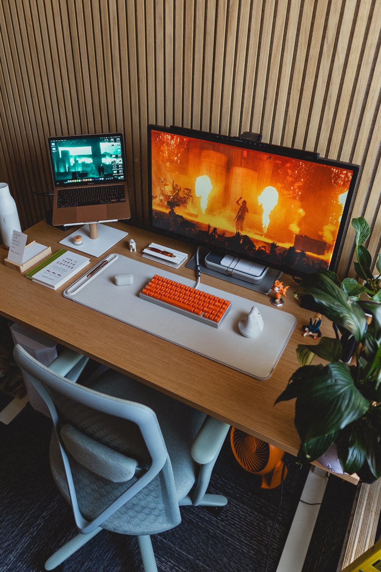  A desk setup with a Branch Verve chair, an M1 MacBook Air on an Ugmonk stand, an HP Pavilion monitor displaying a concert scene, a KMG B65 keyboard with DCX Topaz keycaps, and plants adding a natural touch to the workspace