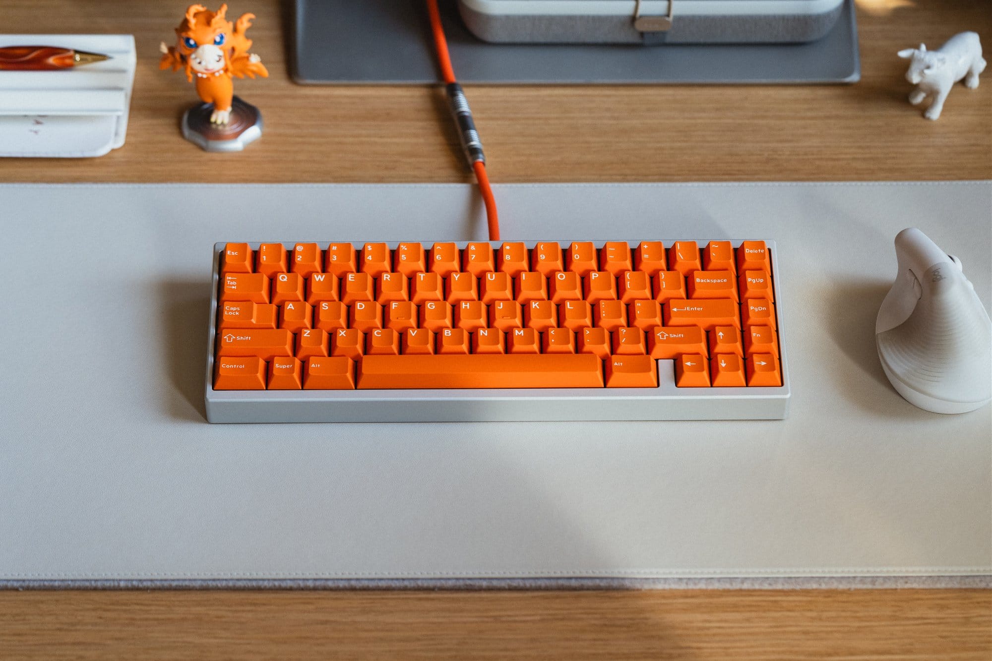 A desk setup showing a KMG B65 keyboard with bright orange DCX Topaz keycaps on an Orbitkey Slim desk mat, alongside a white Logitech Lift mouse