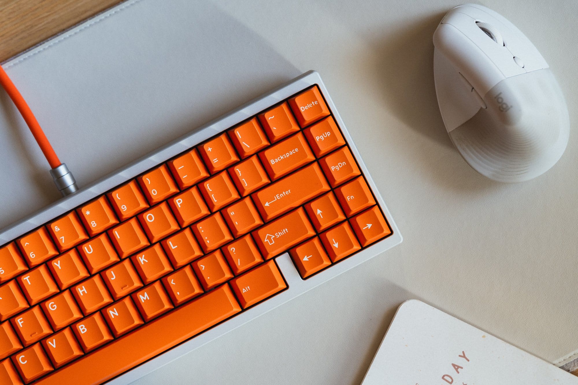 A close-up of a KMG B65 keyboard with bright orange DCX Topaz keycaps next to a white Logitech Lift mouse, placed on a light grey Orbitkey Slim desk mat