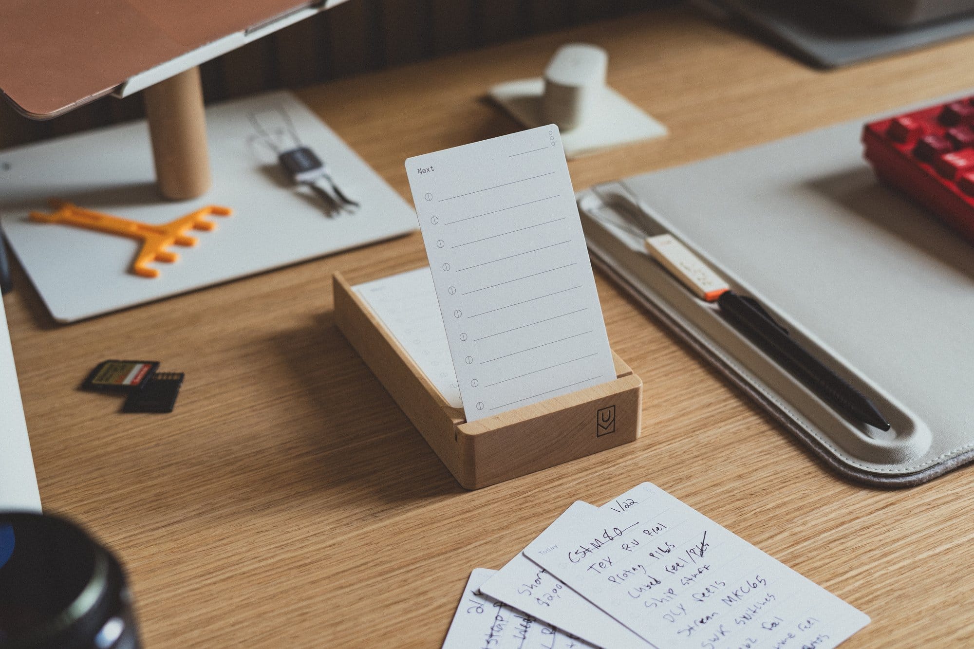 A wooden Ugmonk desk organiser holding to-do cards, surrounded by scattered handwritten notes and stationery on a wooden desk