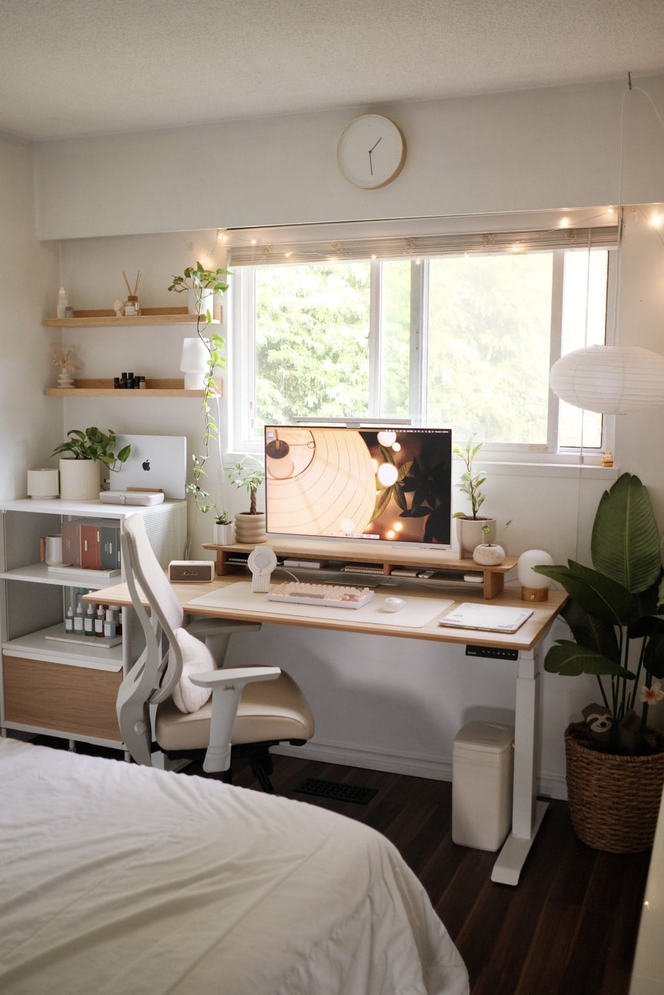 A bedroom workspace with a Samsung monitor on a Branch standing desk, a Keychron Q1 V2 keyboard, and a MacBook Pro on a shelf by the window