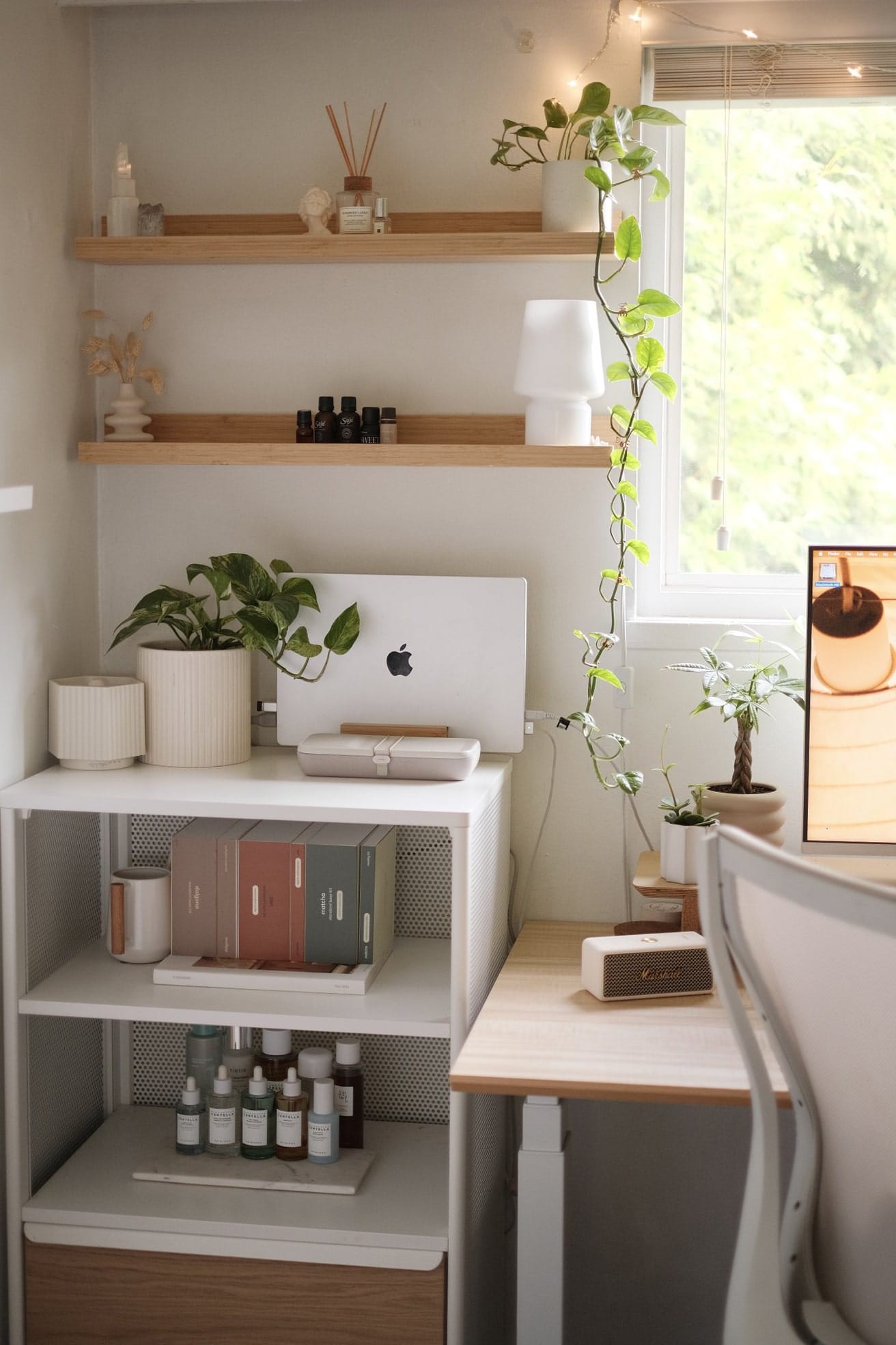 A MacBook Pro 16-inch 2021 on a white side cabinet with a vertical bamboo laptop stand, potted plants, and wooden shelves by a window