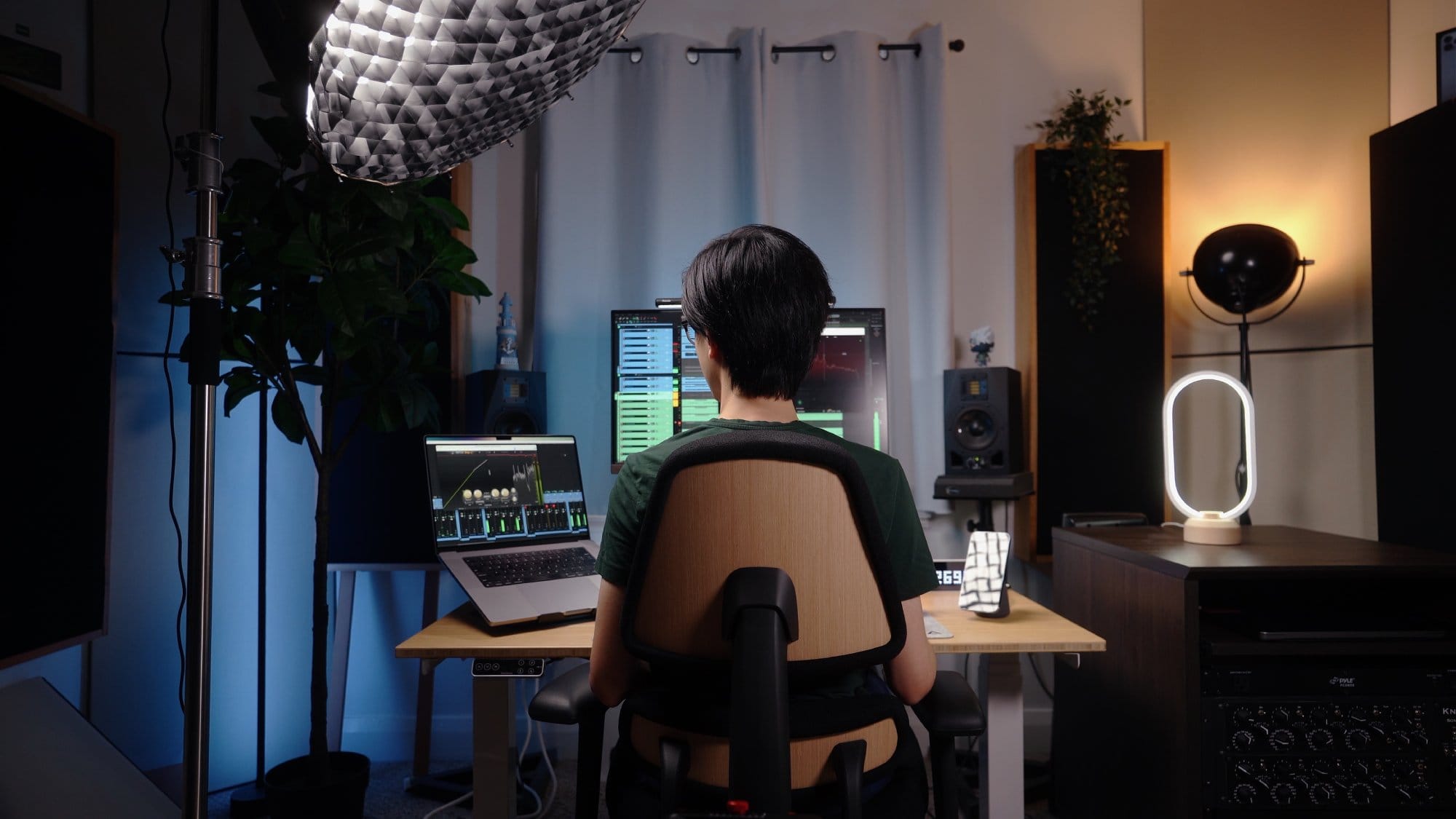 A person sitting on an Anthros Chair, working at a desk setup with an M1 Max MacBook Pro and a BenQ EW2780U monitor, surrounded by tech accessories like Adam A5X speakers and a Belkin 2-in-1 charging dock