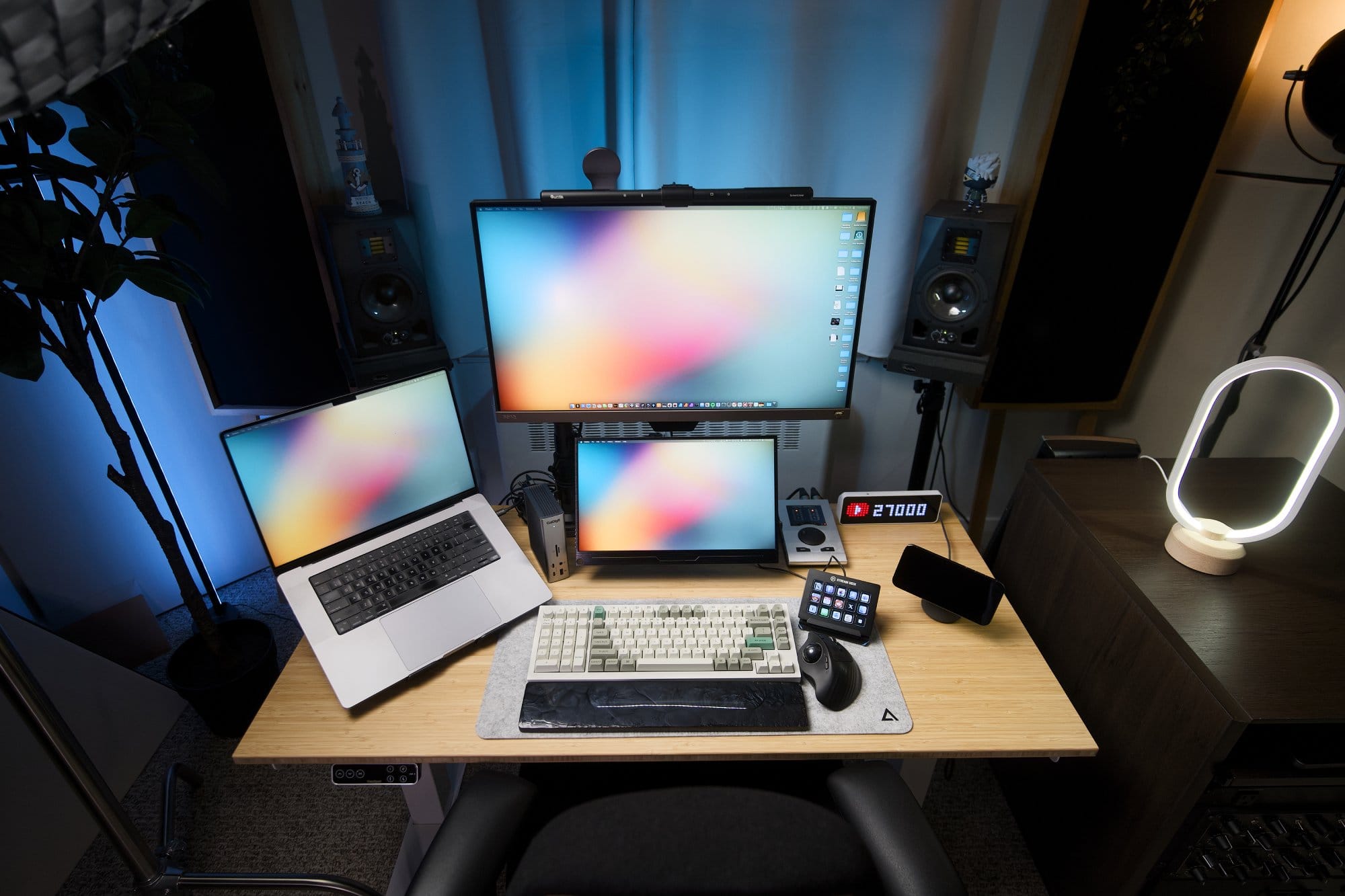 A top-down view of a desk setup featuring an Anthros Chair, a BenQ EW2780U monitor with a Quintis ScreenLinear Light Bar, an M1 Max MacBook Pro, a Keychron Q12 Max keyboard on a DeltaHub Felt Desk Mat, a ProtoArc EM01 trackball mouse, an Ulanzi Smart Pixel Clock, a Stream Deck Classic, and decorative ambient lighting