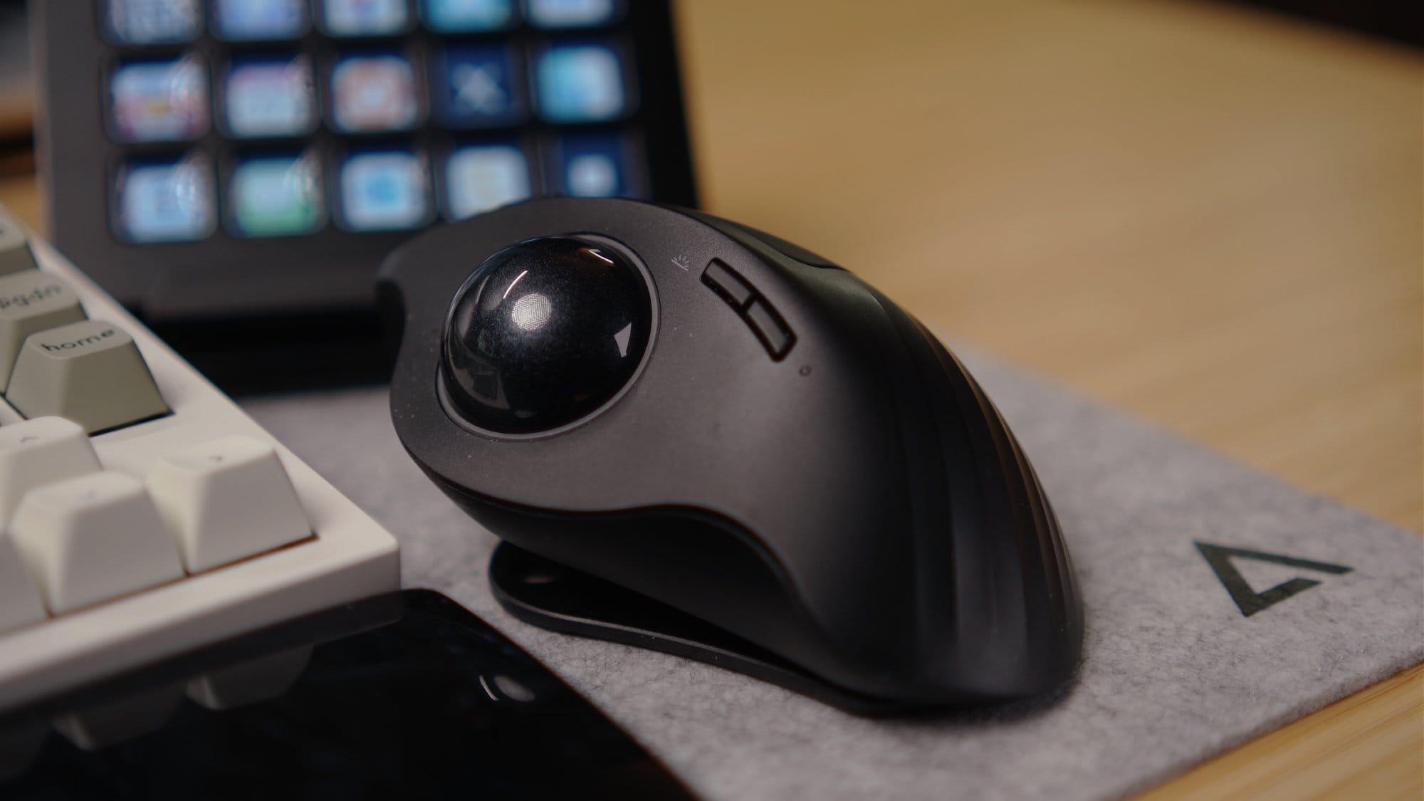 A close-up of a ProtoArc EM01 trackball mouse placed on a DeltaHub felt desk mat, with part of a Keychron Q12 Max keyboard and a Stream Deck visible in the background