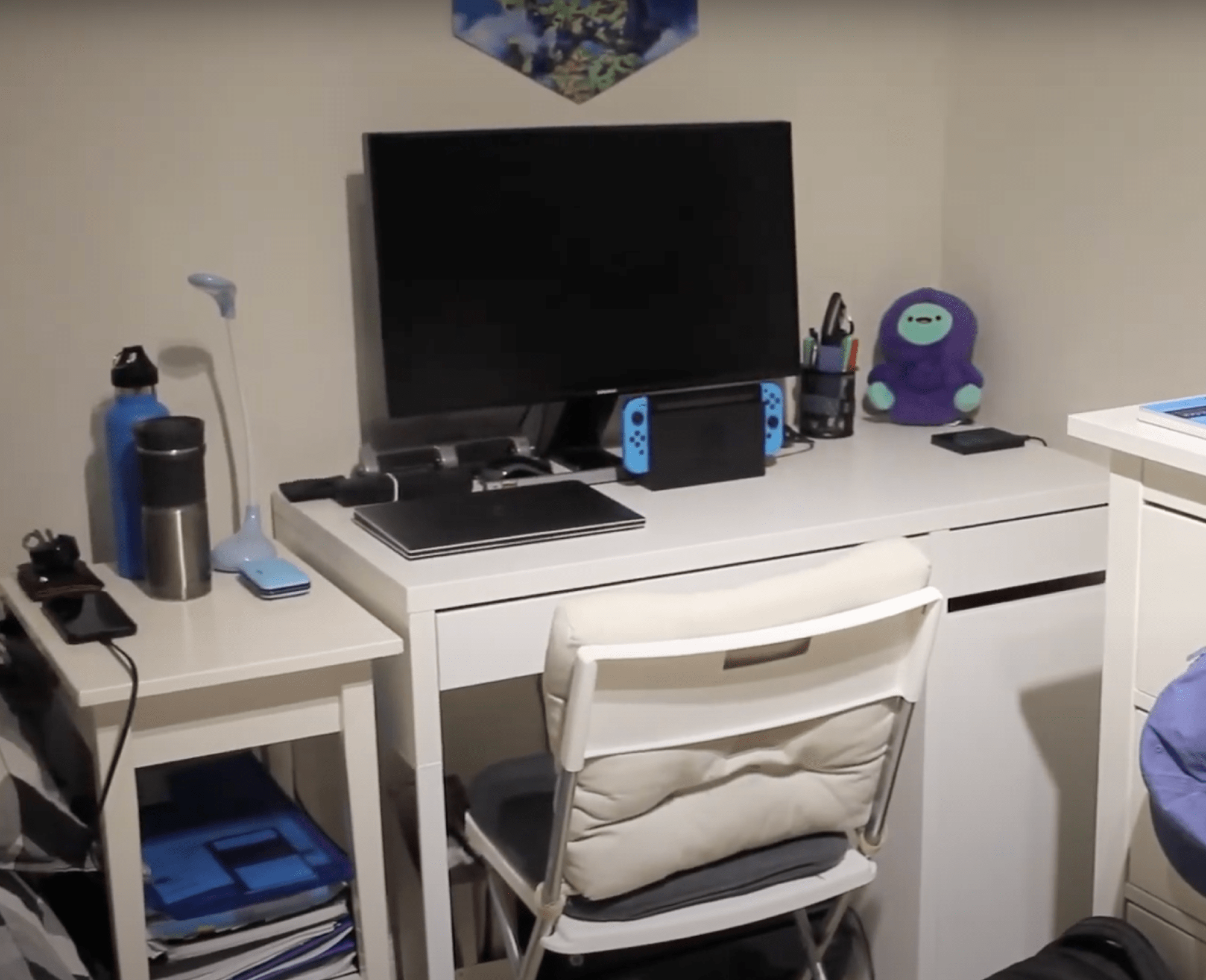 A compact desk setup with a monitor, a Nintendo Switch, a laptop, a white folding chair with cushions, and a small side table holding a water bottle, thermos, and phone