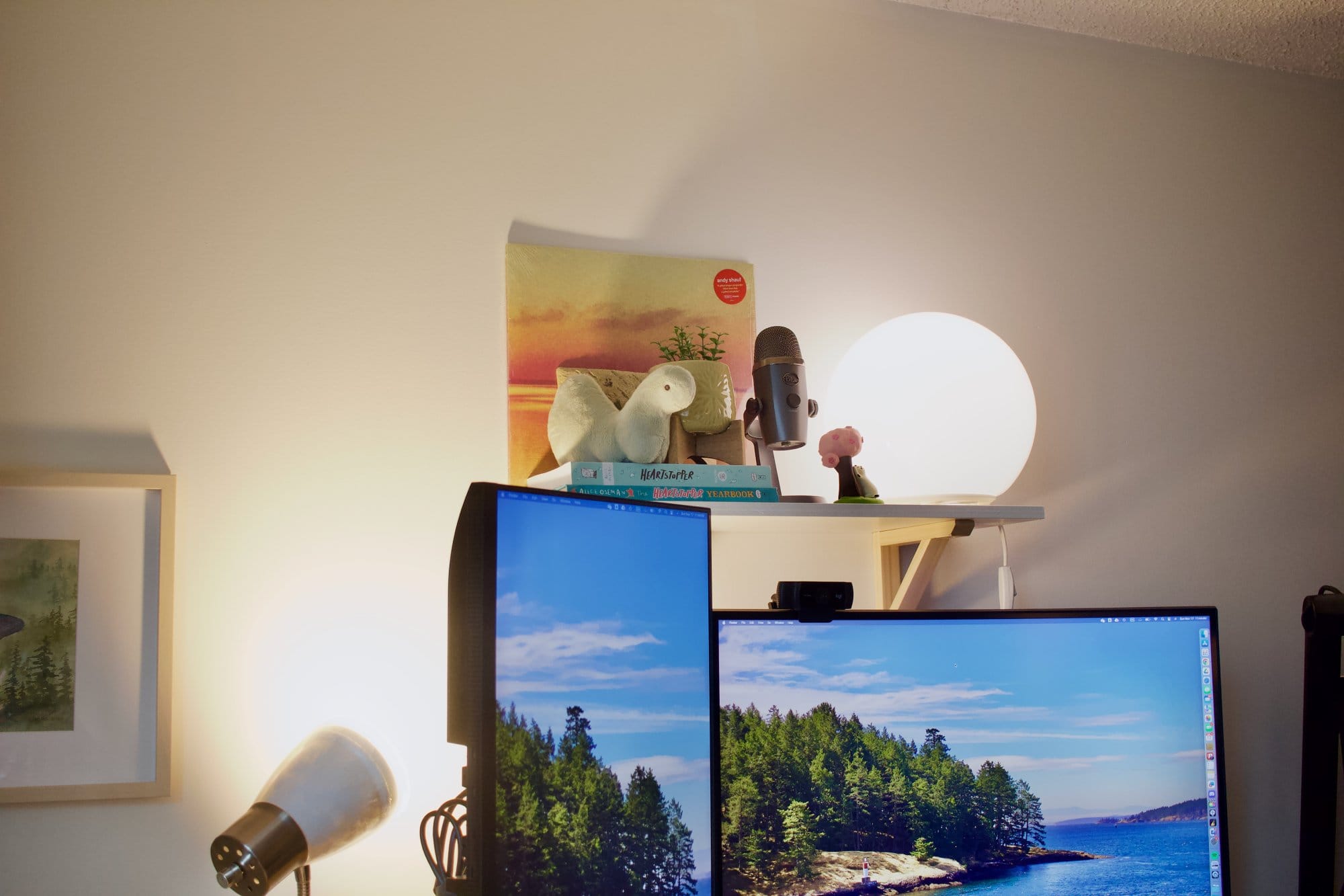 A desk shelf holding a Blue Yeti Nano microphone, books, a stuffed toy, a potted plant, and a glowing spherical lamp, positioned above dual monitors