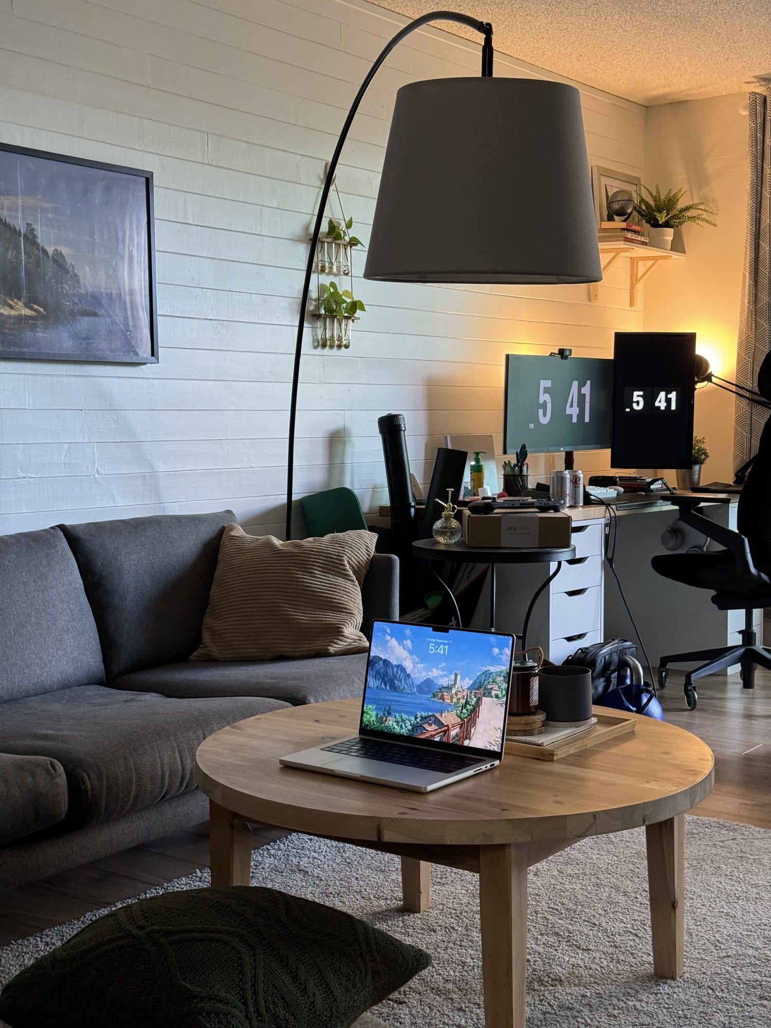 A living room with a wooden coffee table holding a MacBook, a grey sofa with cushions, a large floor lamp, and a desk setup in the background with dual monitors and a black office chair